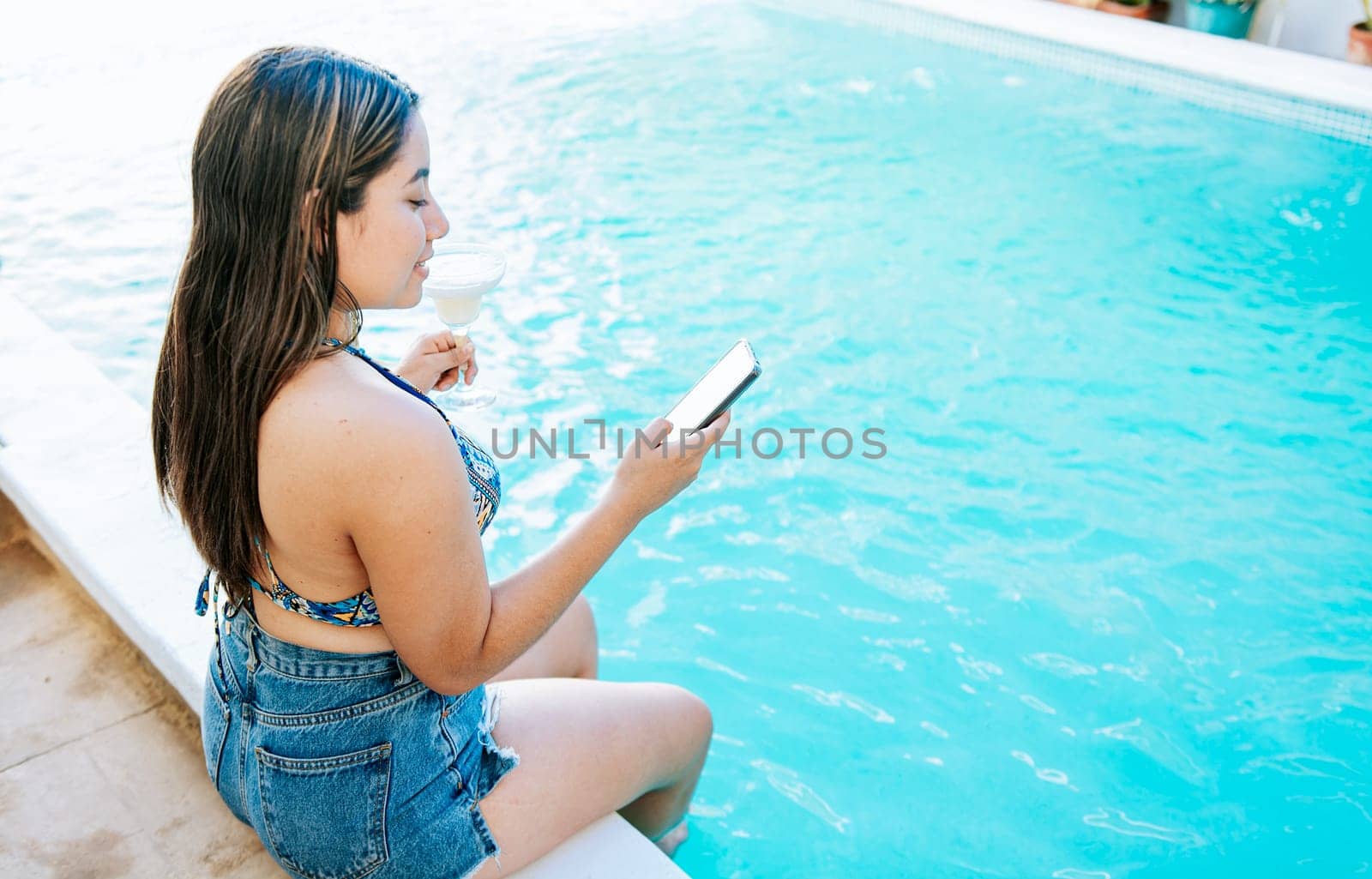 Girl sitting on the edge of the swimming pool holding drink and checking cell phone. Smiling girl on vacation in swimming pool checking cell phone and holding cocktail by isaiphoto