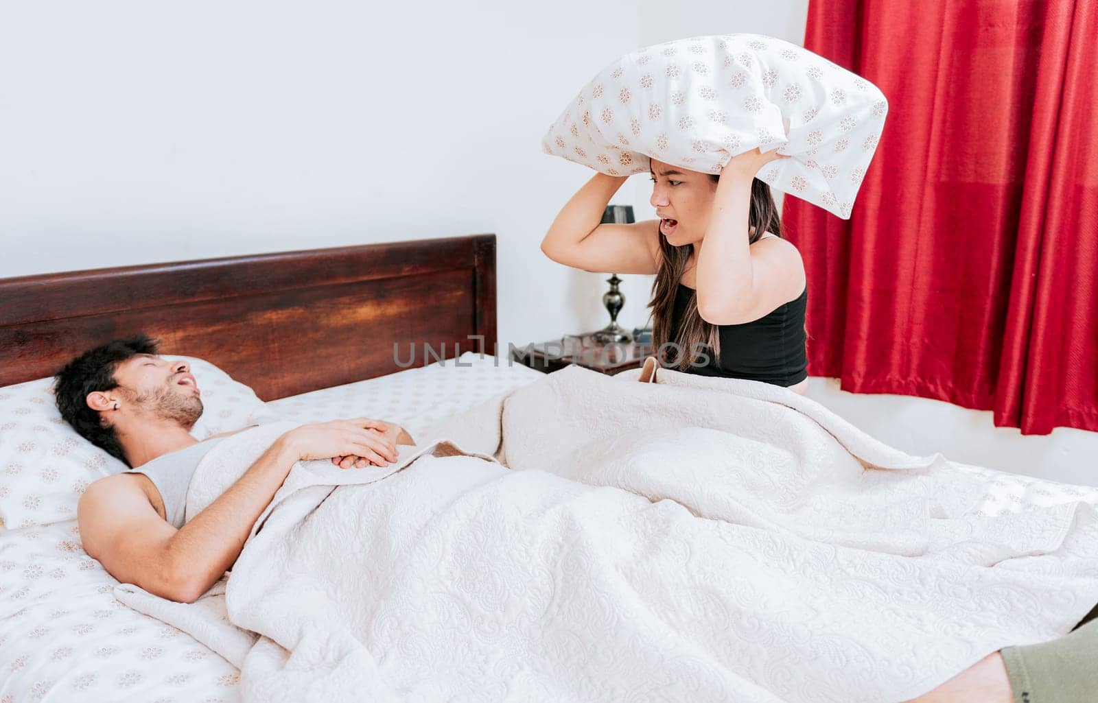 Suffering wife covering her ears while her husband snores. Angry woman holding pillow and snoring husband, Snoring man in bedroom and wife covers her ears with pillow by isaiphoto