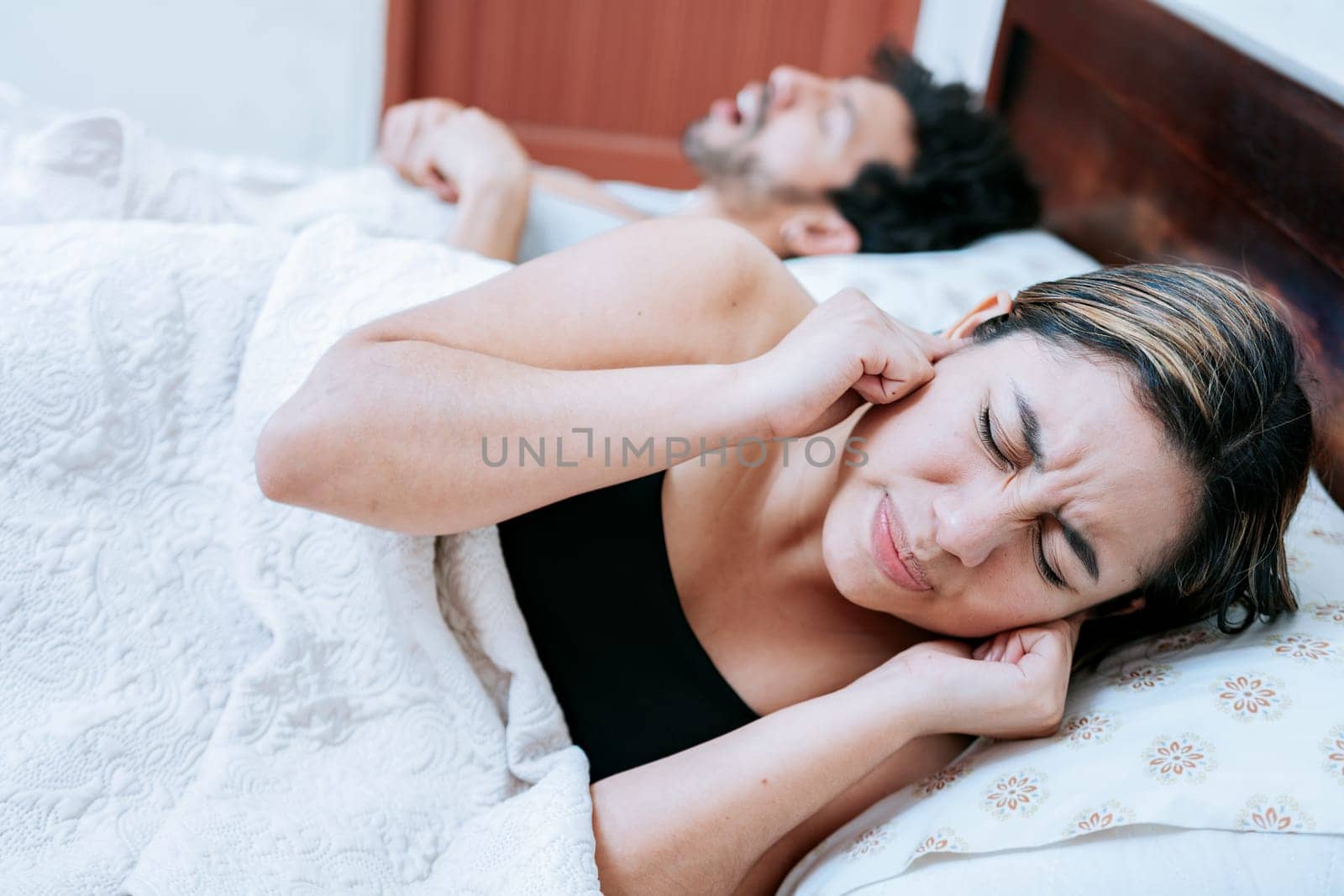 Wife suffering from her sleeping husband snoring. Sleep apnea concept, man snoring in bedroom and wife covering her ears by isaiphoto