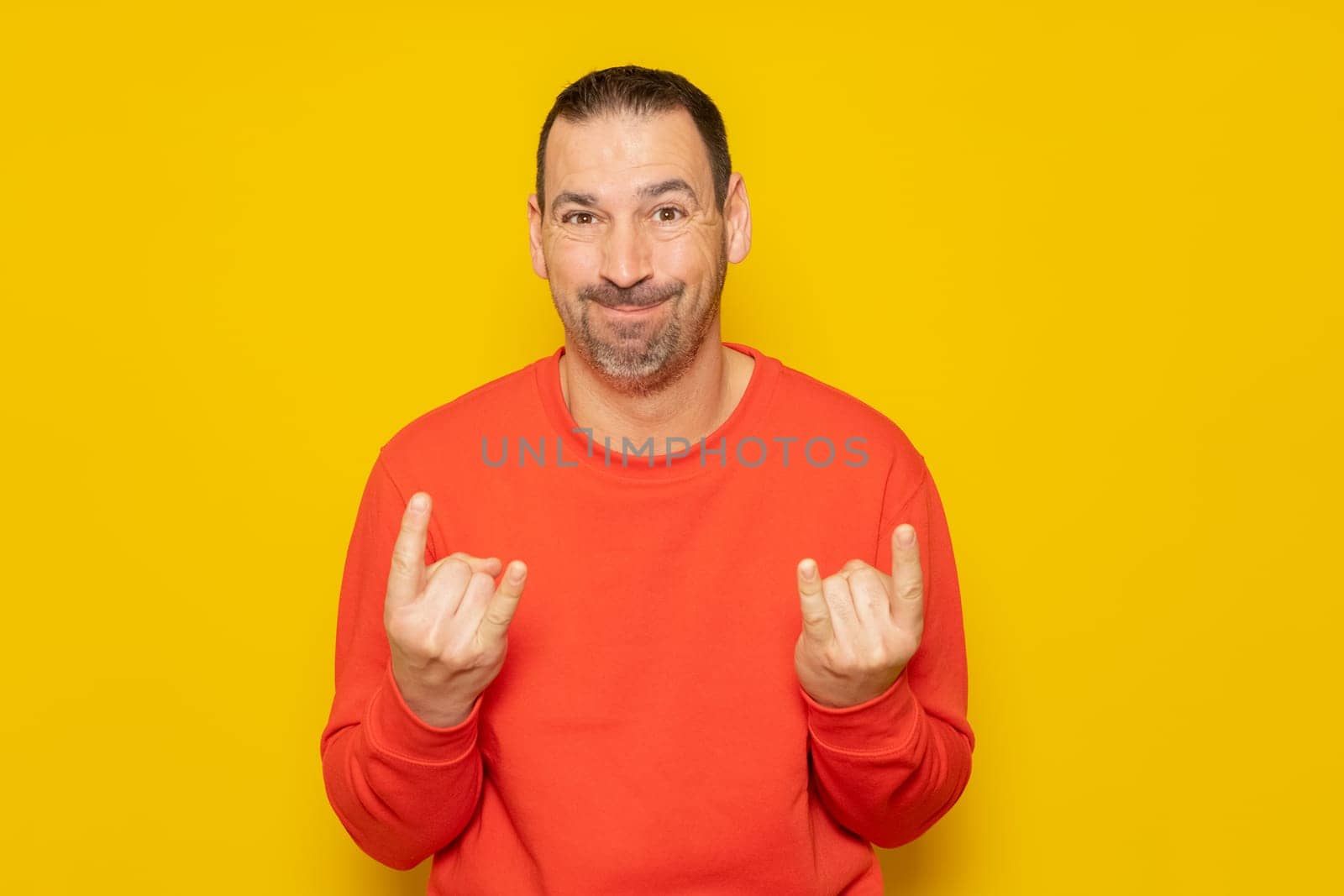 Hispanic man with a beard wearing a red pullover making the rock gesture with arms close to the body isolated on yellow studio background, isolated on yellow background.