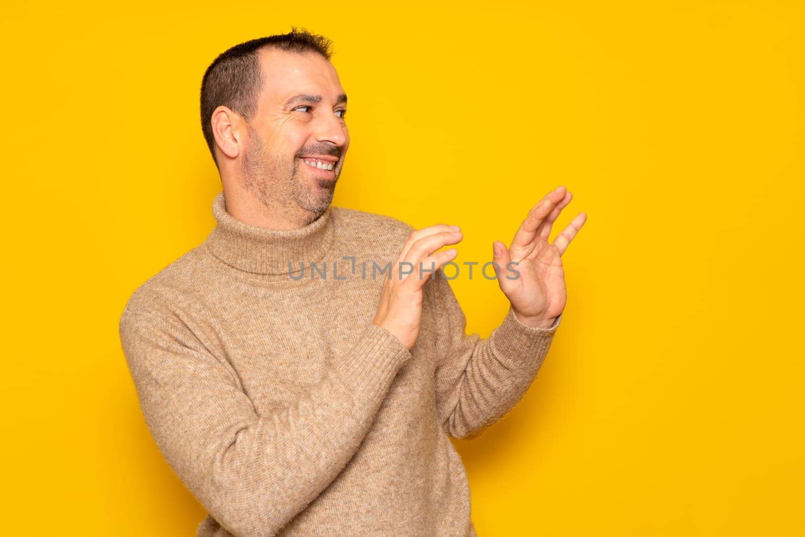 Bearded hispanic man in a turtleneck ducking to the side in embarrassment, tries to push away from something or someone with his hands. Isolated on yellow background