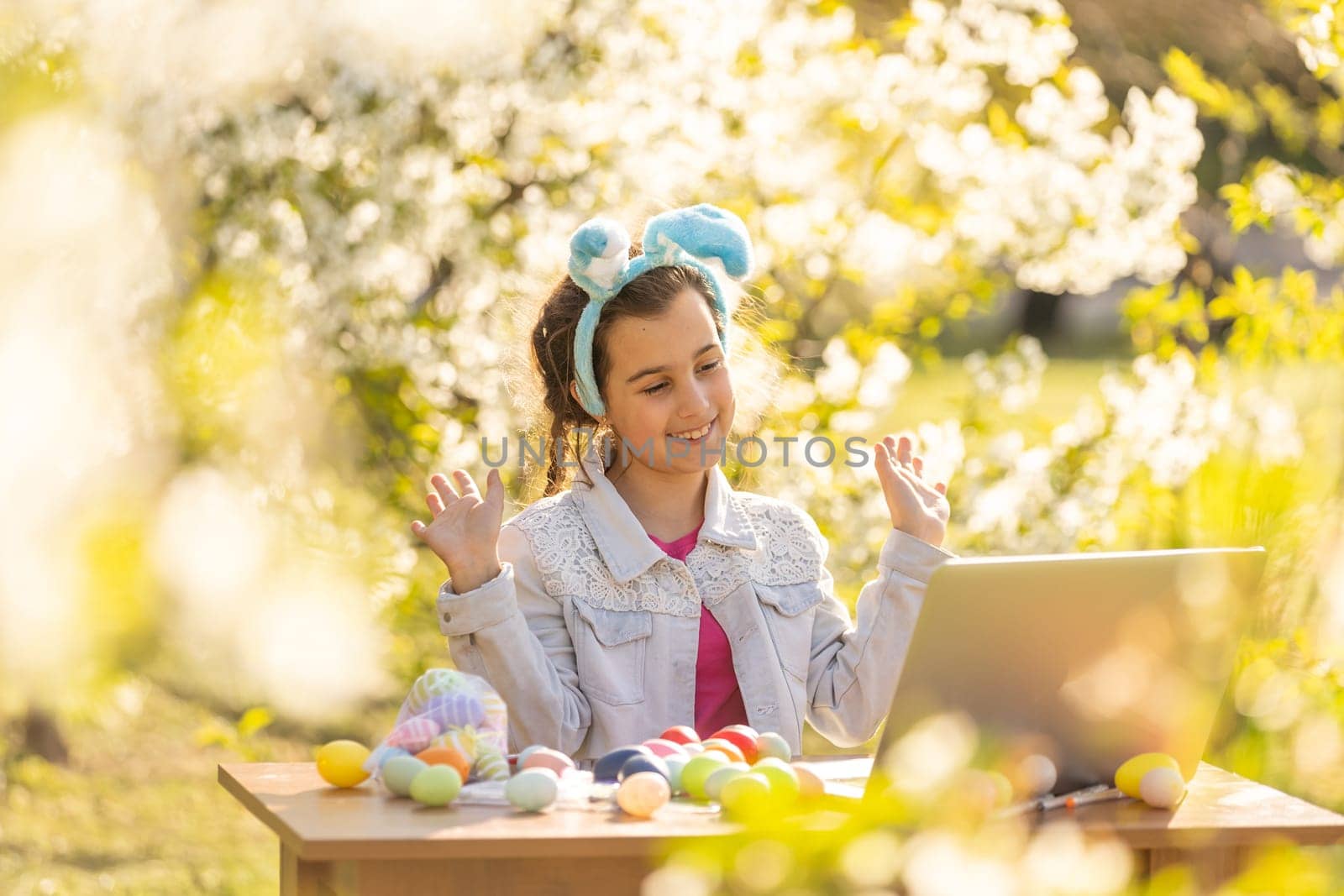 happy teen girl wear bunny ears paints Easter eggs with a laptop in the garden by Andelov13