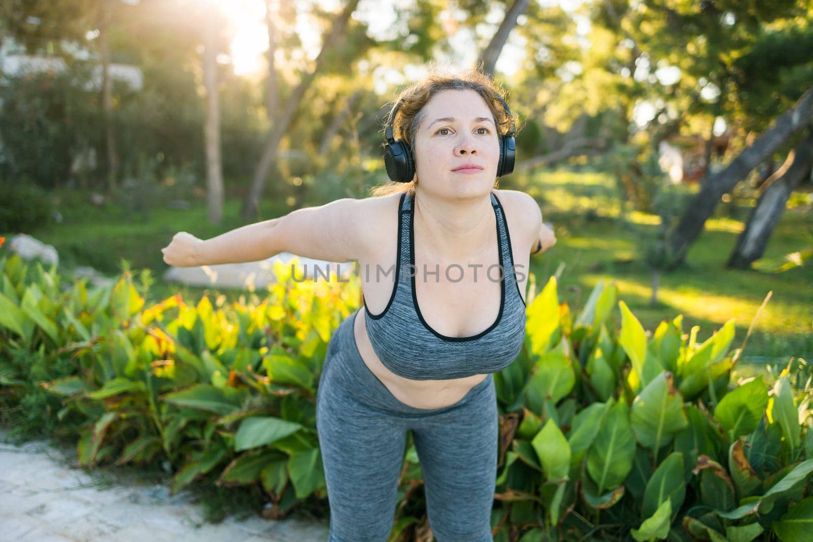 Young pretty smiling plus size woman in sporty top and leggings doing sport fitness and listen to music with headphones in summer morning outdoor copy space by Satura86