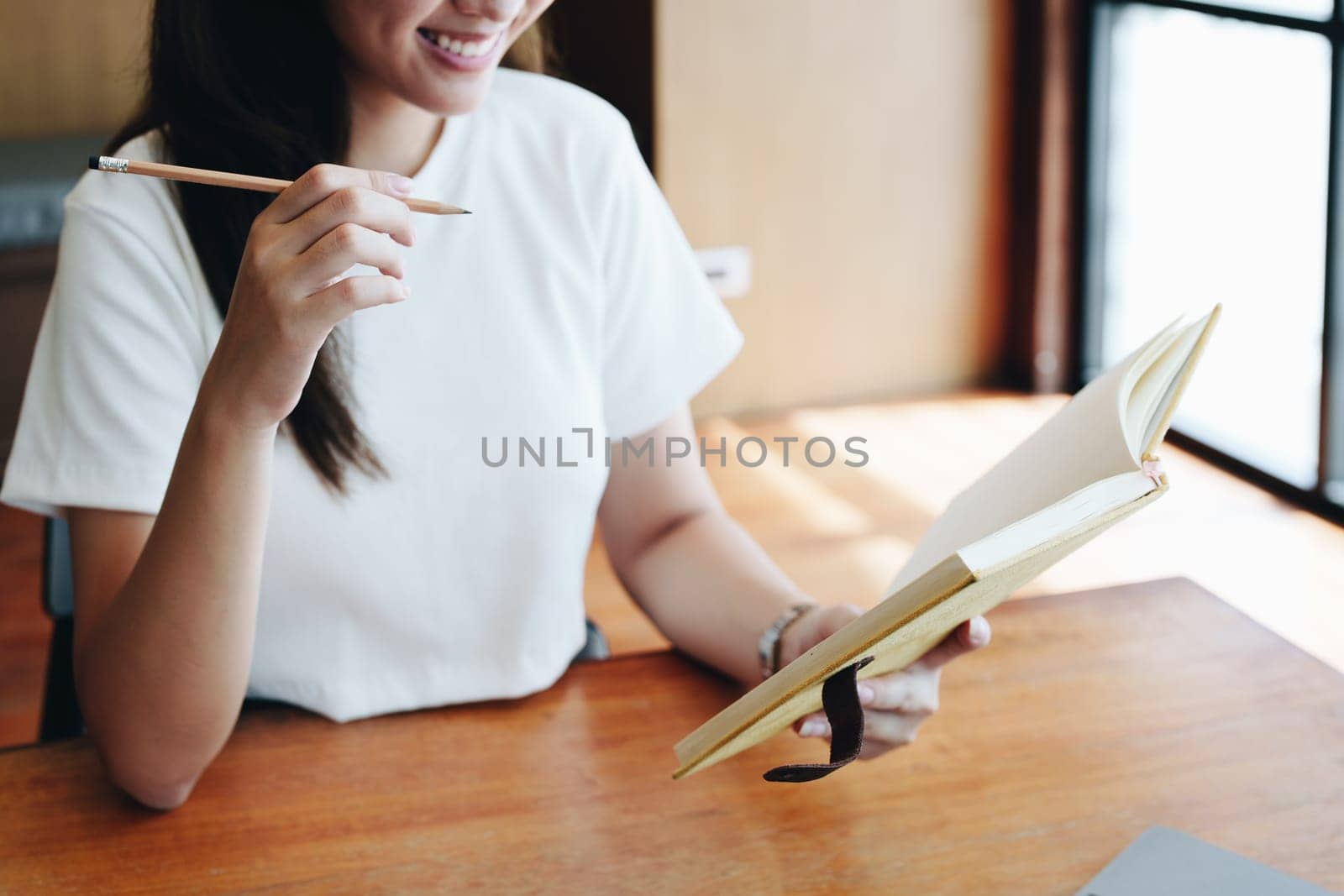 Portrait of a teenage Asian woman using a computer, wearing headphones and using a notebook to study online via video conferencing on a wooden desk in library by Manastrong