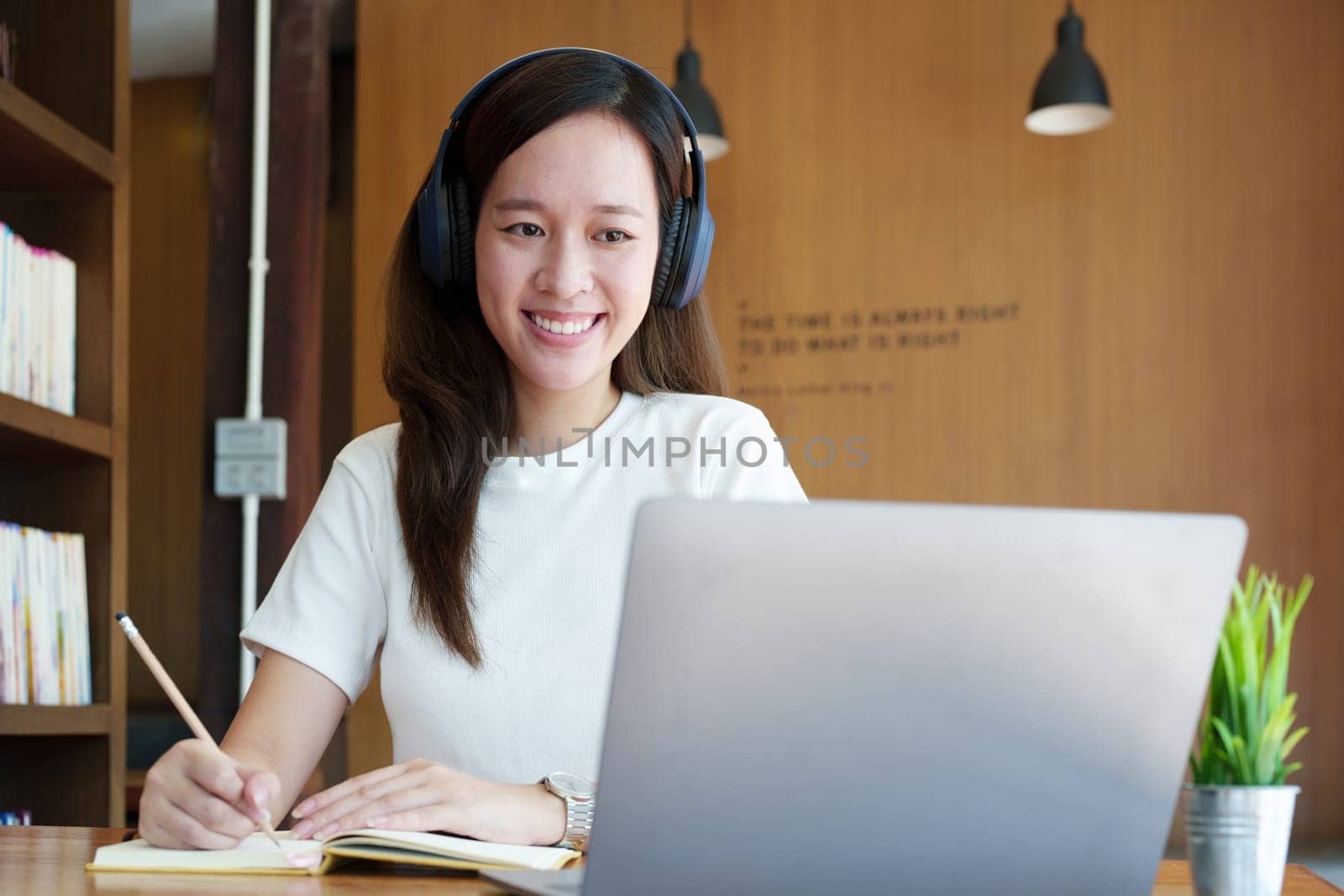 Portrait of a teenage Asian woman using a computer, wearing headphones and using a notebook to study online via video conferencing on a wooden desk in library by Manastrong
