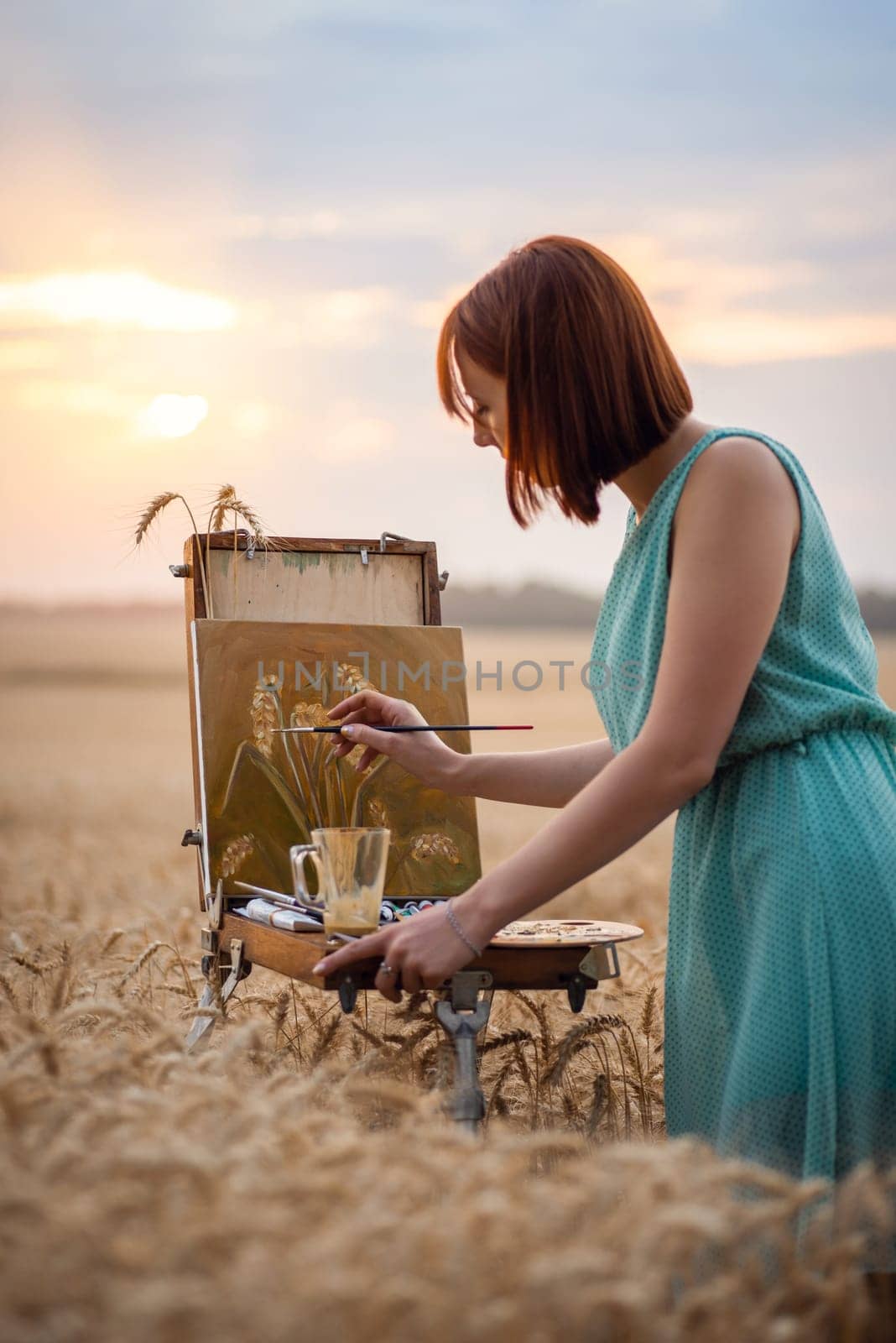 Girl artist painting ears of wheat in field of rye at sunset