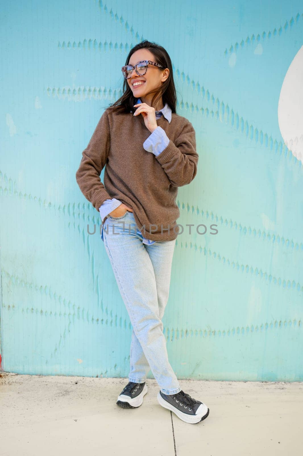 Happy woman in glasses outdoor on blue color background. Positive people concept. Smiling girl looking away, hands in pocket, dressed sweater and jeans. Vertical full length