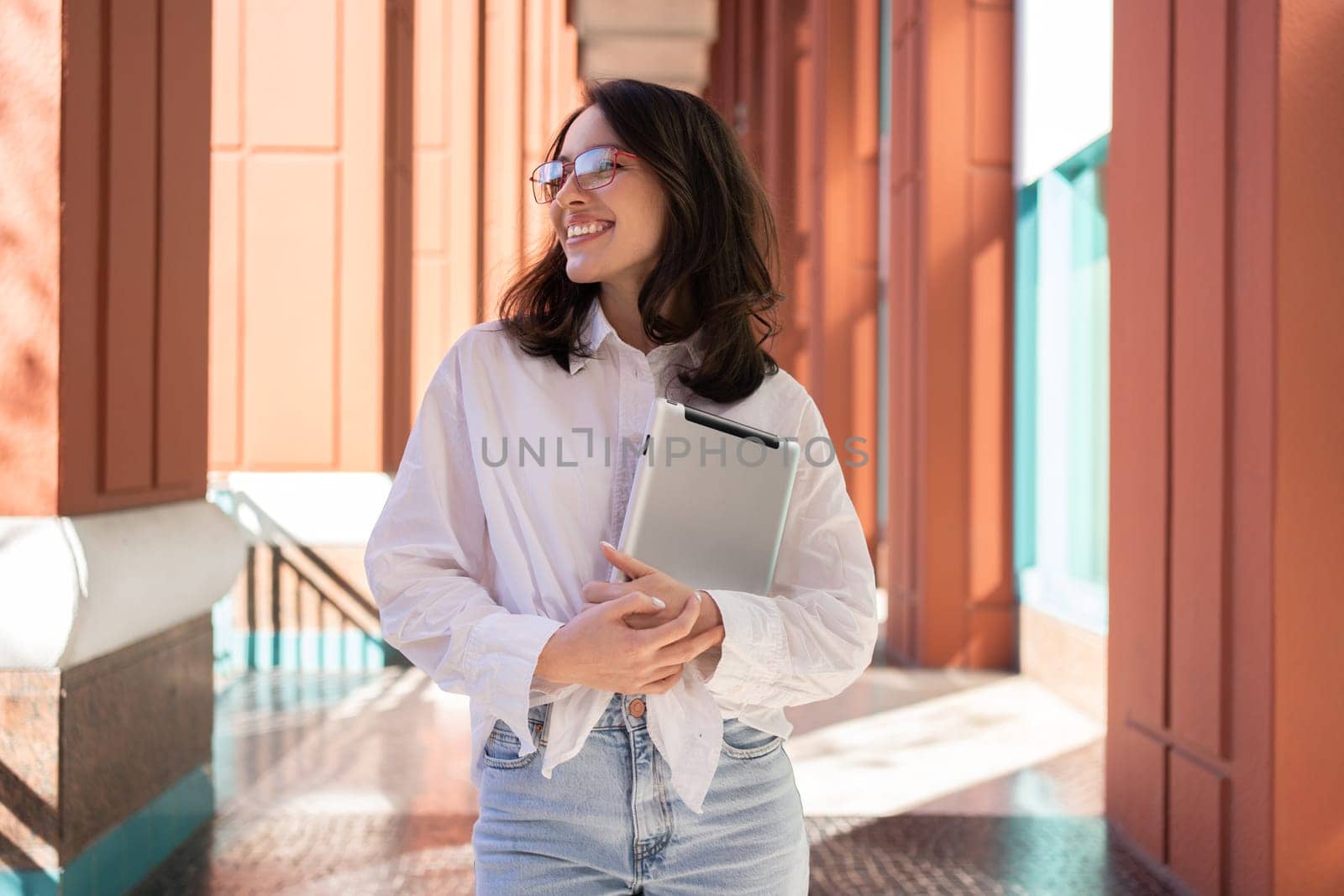 Businesswoman in glasses looking away, smiling and holding digital tablet, standing outdoor. Female freelancer holding tablet while walk outside