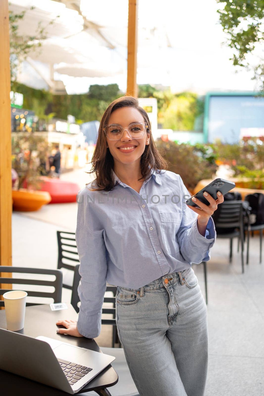 Freelance worker doing remote work using smartphone and laptop, looking at camera and smile. Female freelancer with mobile phone standing in outdoor cafe holding phone in hand. Vertical shoot