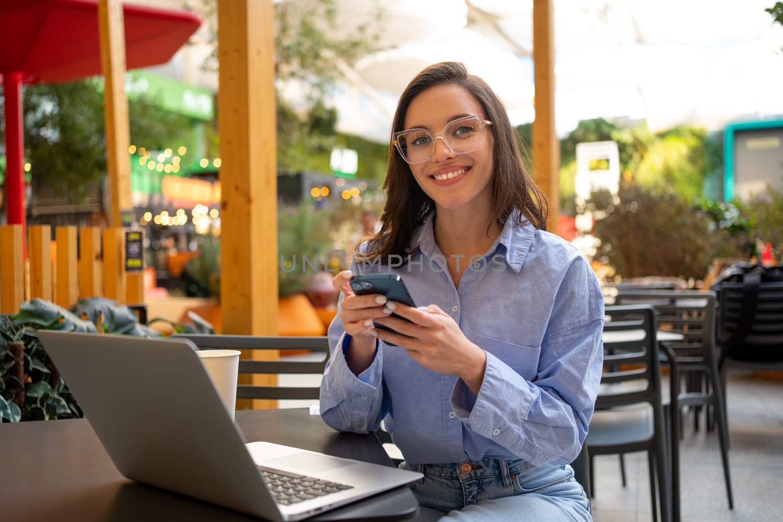 Freelance worker doing remote work using smartphone internet. looking at camera and smile. Business woman with mobile phone in outdoor cafeteria, restaurant on social media app online.