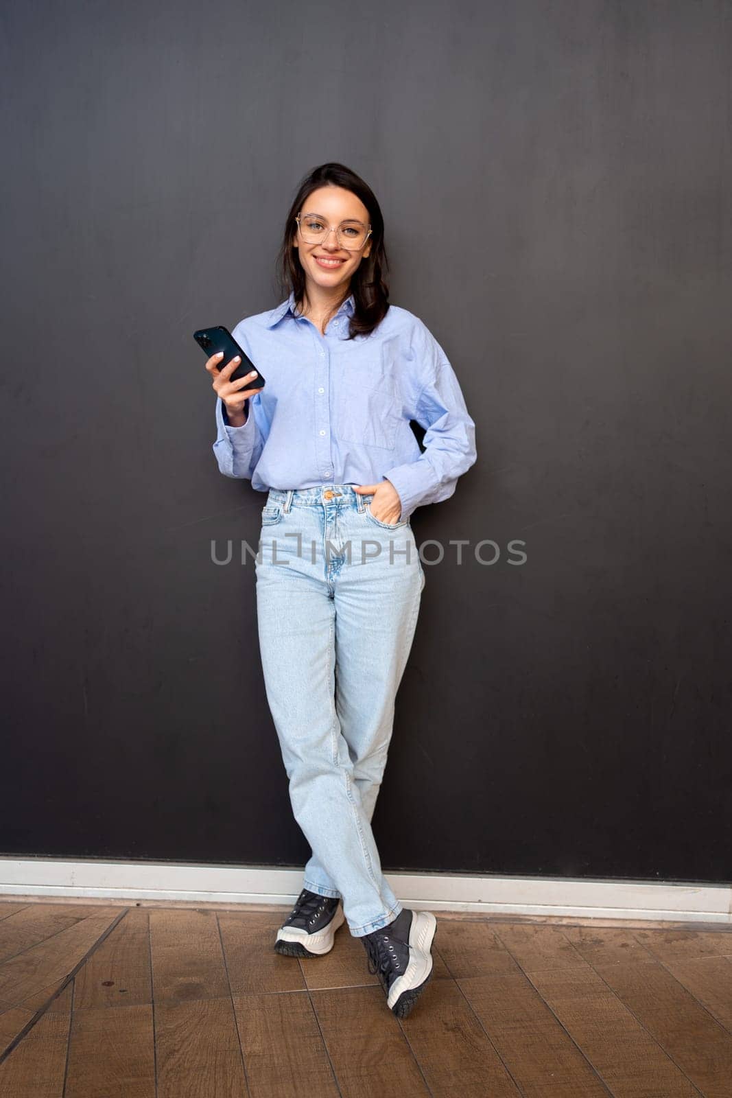Happy hipster girl wear glasses, types text message on mobile phone, enjoys online communication, types feedback on black studio background. Full length, looking at camera and smile