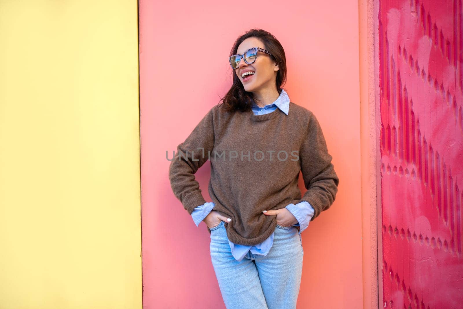 Happy woman in glasses outdoor on yellow and pink color background. Positive people concept. Smiling girl looking side, hands in pocket, dressed sweater and jeans