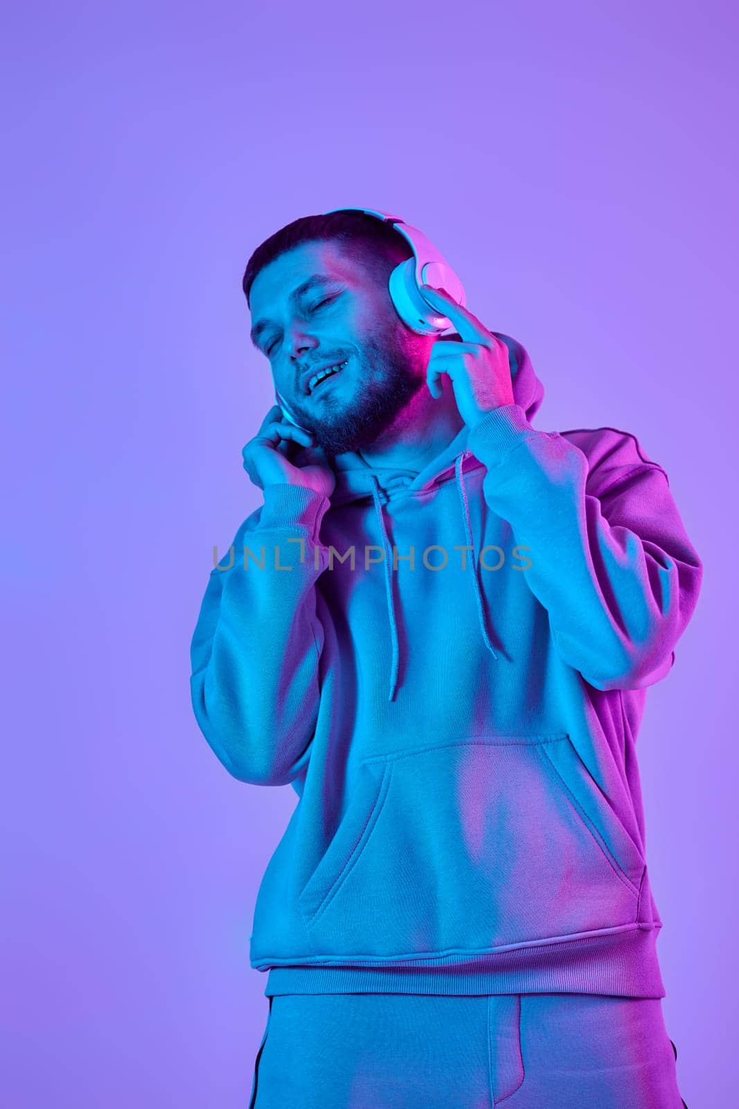 handsome bearded man with headphones in sweatshirt enjoying favorite music on purple neon background. Neon lighting