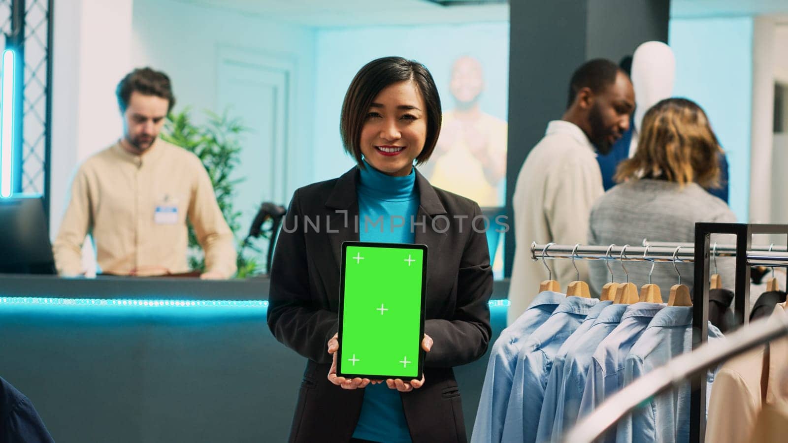 Asian store assistant using greenscreen display on tablet, holding isolated mockup display in clothing store. Cheerful boutique worker showing chroma key copyspace template in retail market.