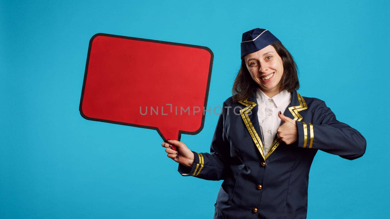 Positive woman holding red speech bubble on camera, working on commercial flights ad in studio. Smiling air hostess showing blank copyspace on carton board, flying industry job.