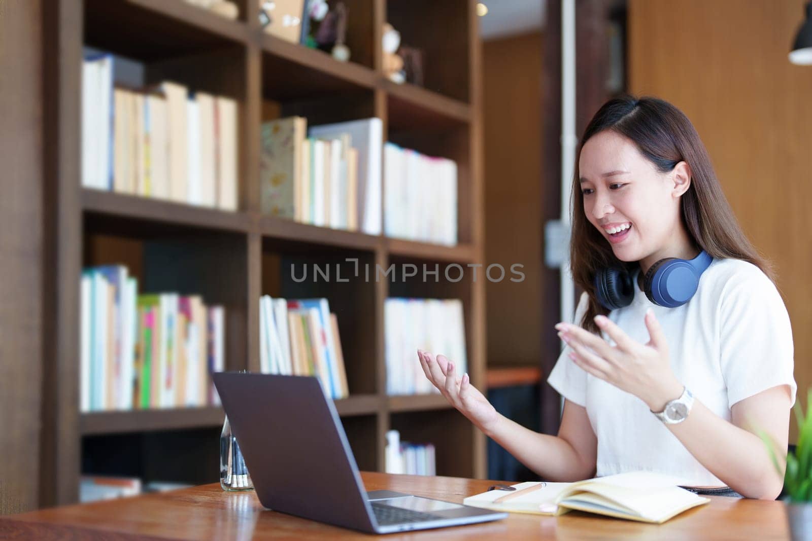 Portrait of a teenage Asian woman using a computer and notebook to study online via video conferencing on a wooden desk in library.