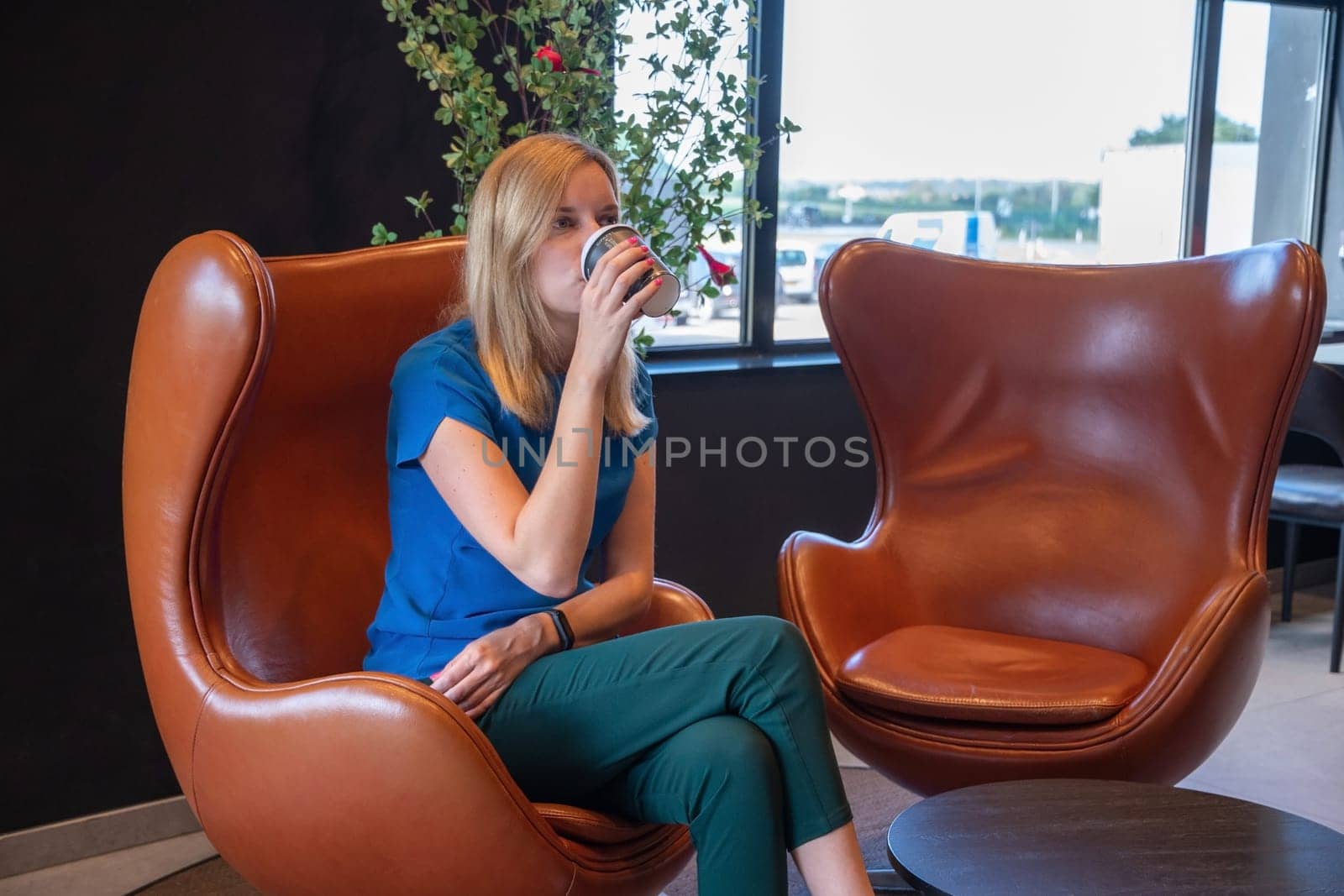 millennial blonde businesswoman sits at a table in a cafe and drinks coffee from a cardboard cup. High quality photo