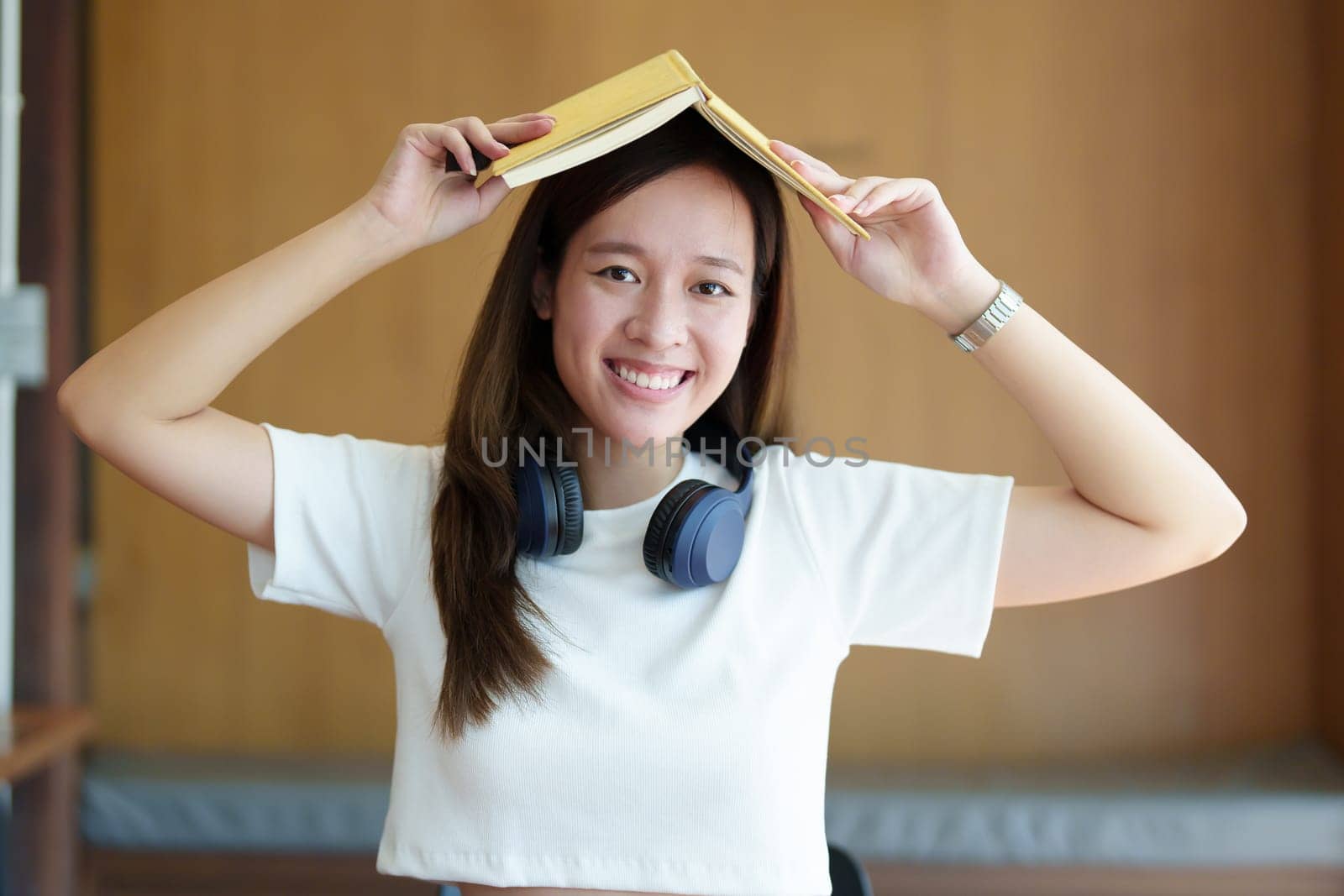 Portrait of a teenage Asian woman wearing headphones and holding a notebook on her head showing a happy face.