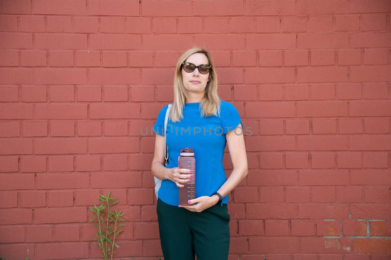 a blonde in a blue blouse against a brick wall drinks water from sports water bottle with time markers, to comply with the drinking regime, High quality photo