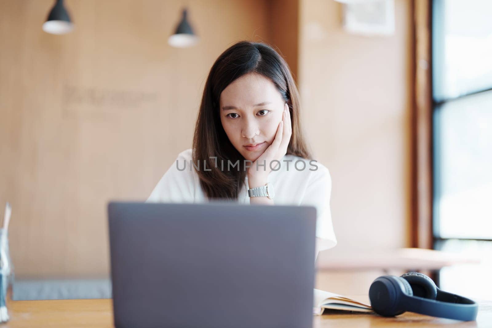 Portrait of a teenage Asian woman using a computer and notebook to study online via video conferencing on a wooden desk in library.