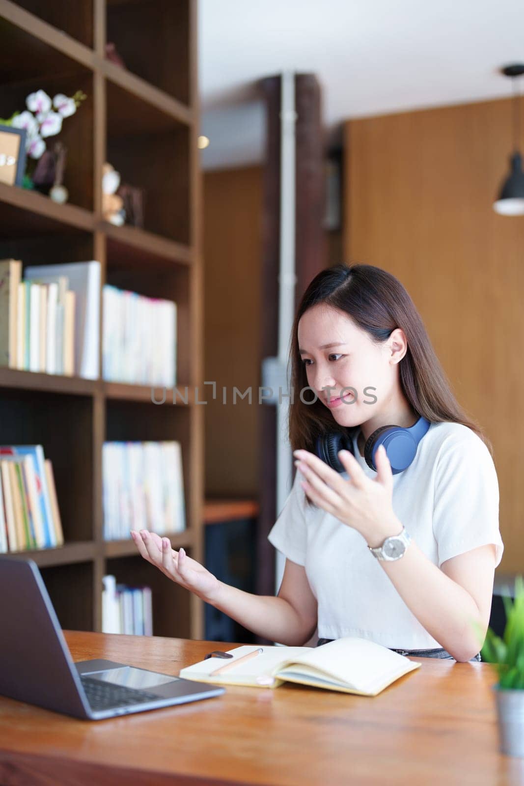 Portrait of a teenage Asian woman using a computer and notebook to study online via video conferencing on a wooden desk in library by Manastrong