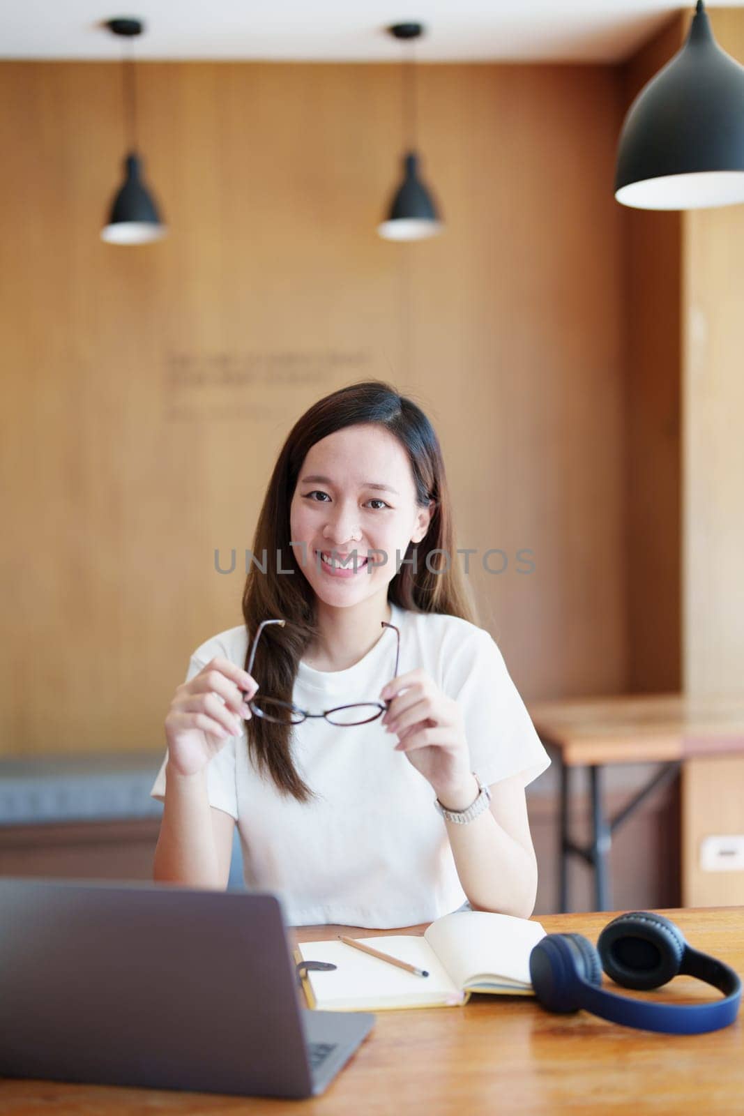 Portrait of a teenage Asian woman using a computer and notebook to study online via video conferencing on a wooden desk in library by Manastrong