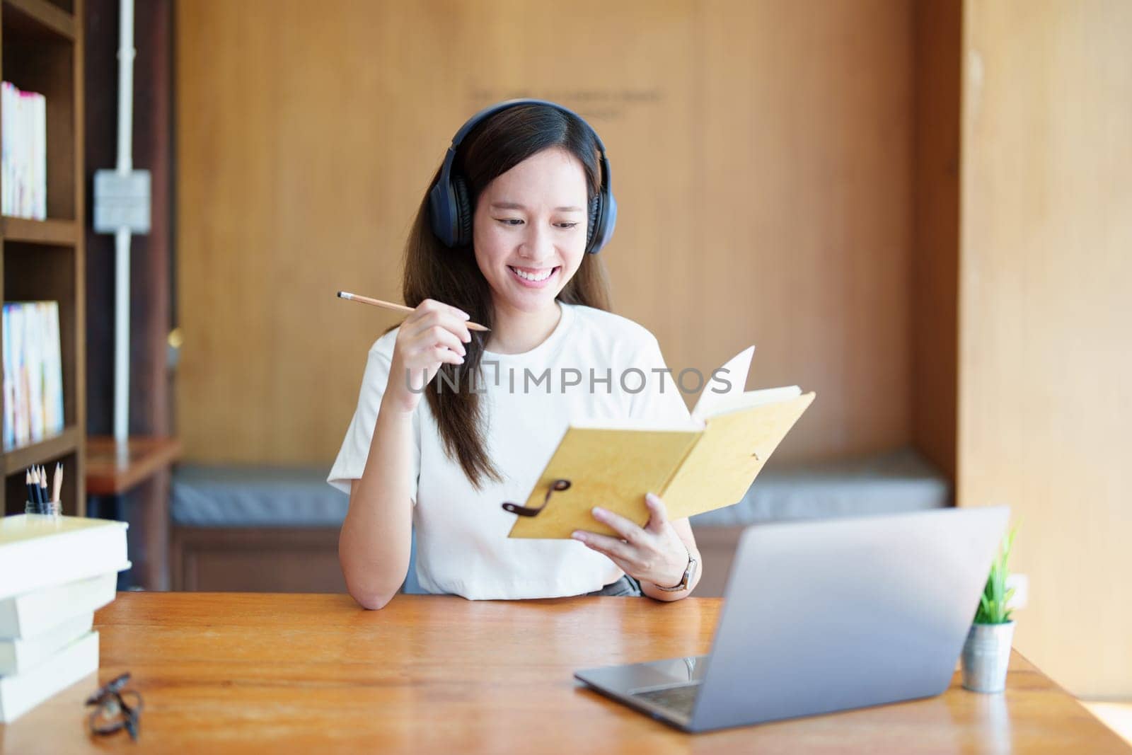Portrait of a teenage Asian woman using a computer, wearing headphones and using a notebook to study online via video conferencing on a wooden desk in library.
