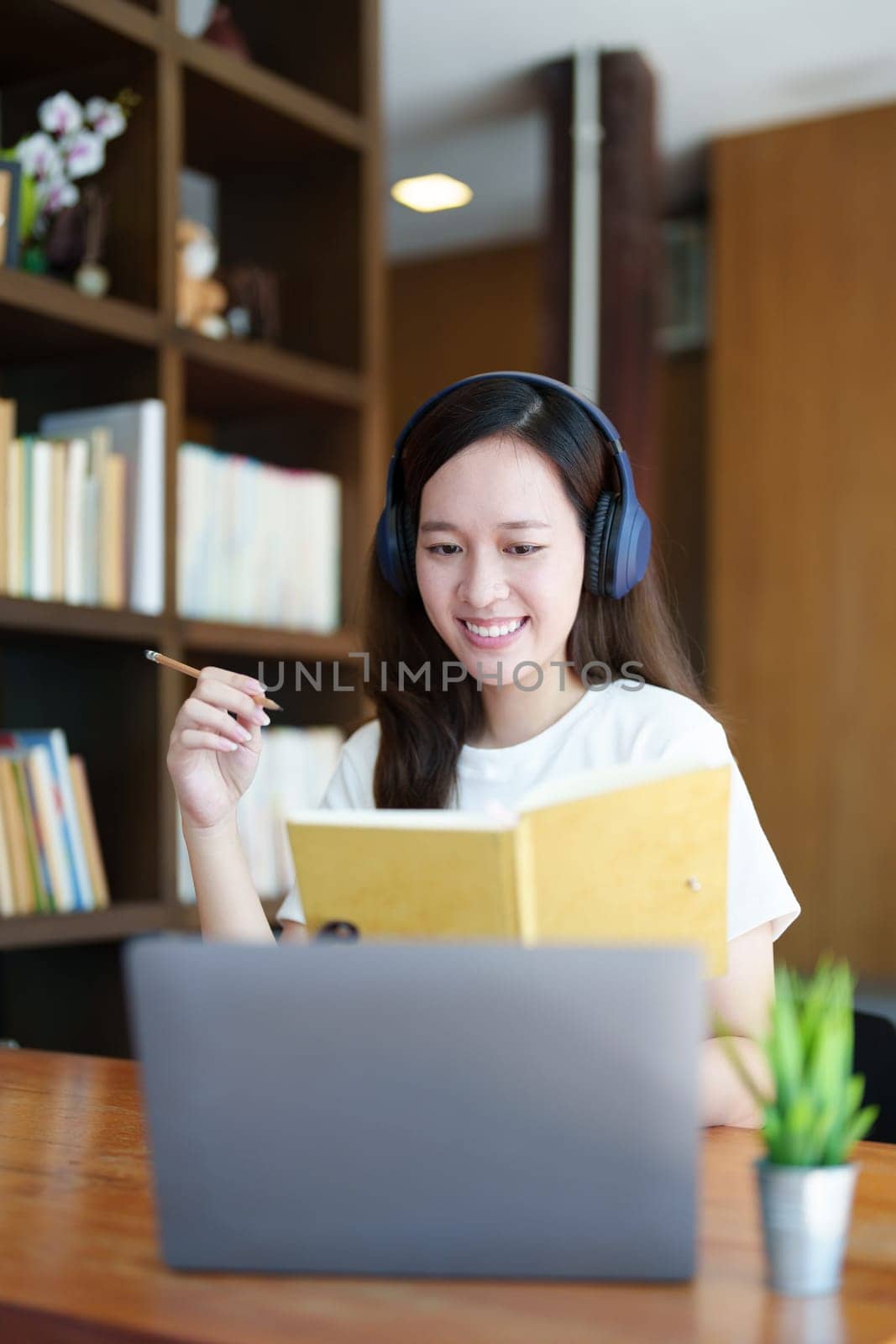 Portrait of a teenage Asian woman using a computer, wearing headphones and using a notebook to study online via video conferencing on a wooden desk in library by Manastrong