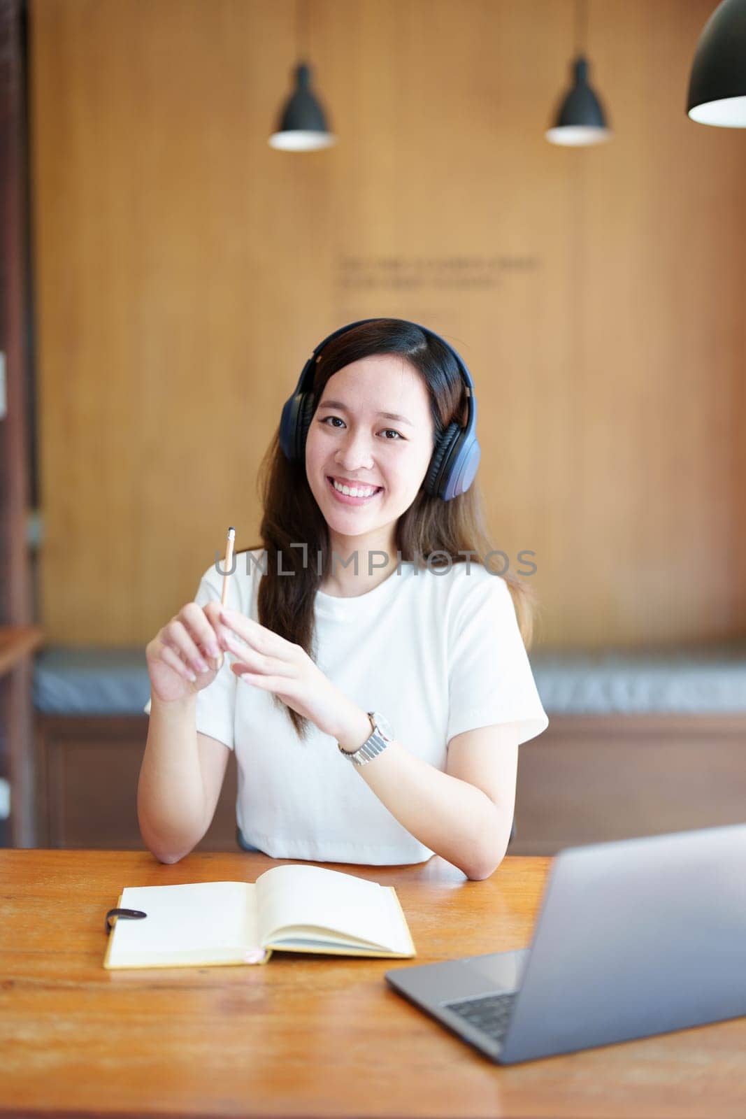 Portrait of a teenage Asian woman using a computer, wearing headphones and using a notebook to study online via video conferencing on a wooden desk in library.