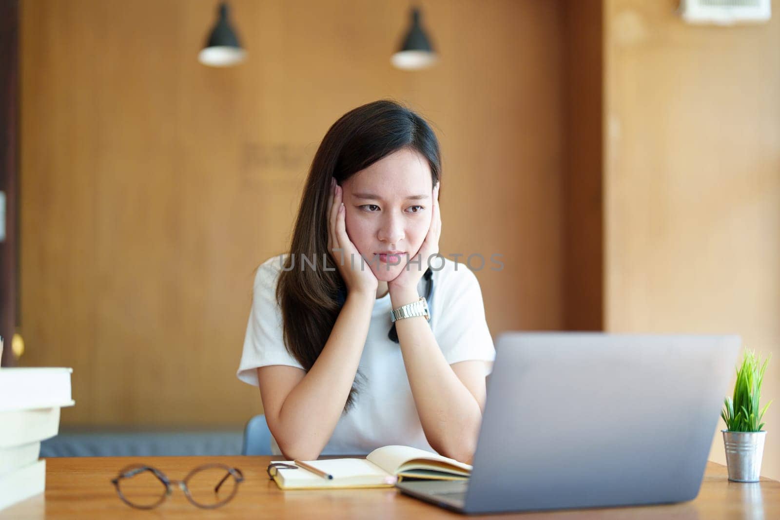 A portrait of a young Asian woman using a computer, wearing headphones and using a notebook to study online shows boredom and pain from video conferencing on a wooden desk in library by Manastrong