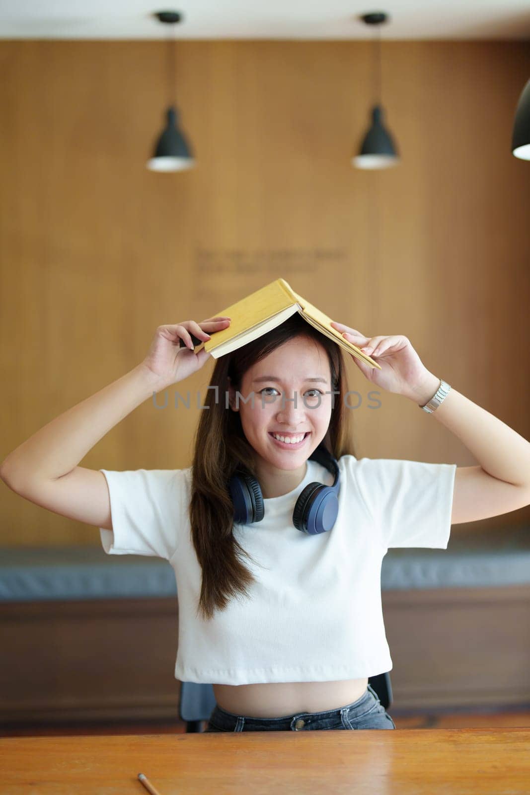 Portrait of a teenage Asian woman wearing headphones and holding a notebook on her head showing a happy face by Manastrong
