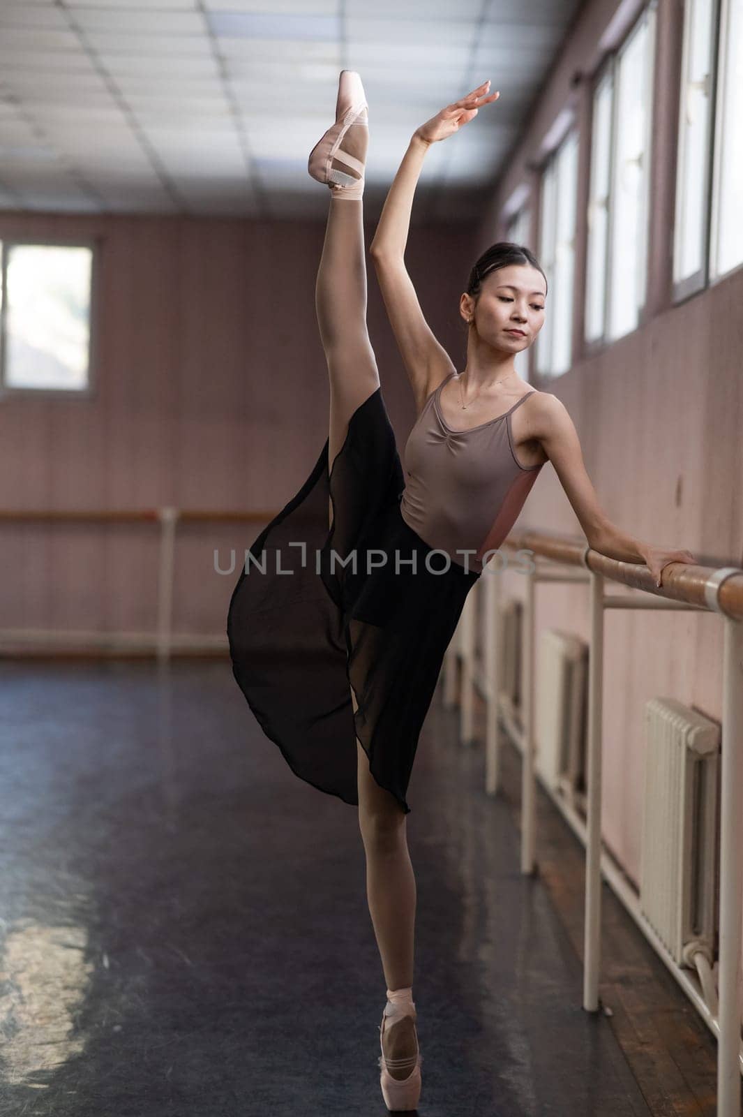 Graceful Asian ballerina in a beige bodysuit and black skirt is rehearsing in a dance class