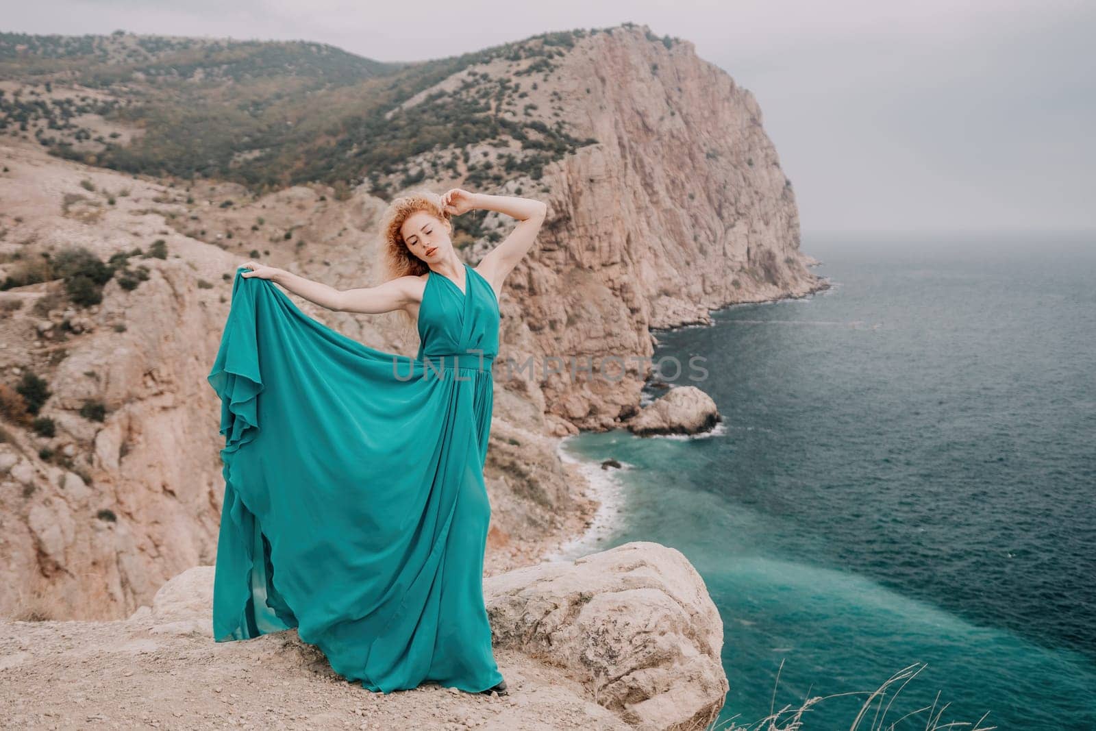 Side view a Young beautiful sensual woman in a mint long dress posing on a volcanic rock high above the sea during sunset. Girl on the nature on overcast sky background. Fashion photo