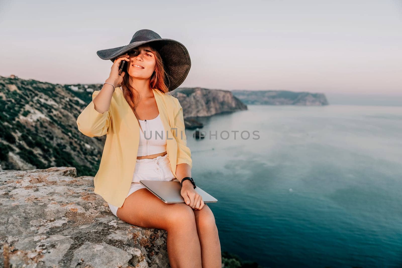 Successful business woman in yellow hat working on laptop by the sea. Pretty lady typing on computer at summer day outdoors. Freelance, travel and holidays concept.
