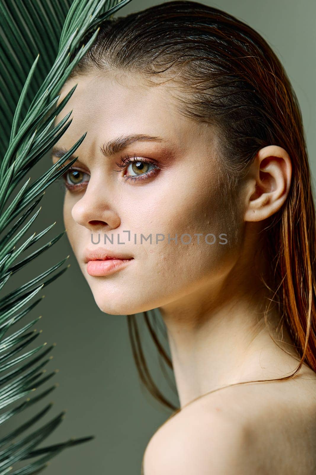 vertical photo, portrait of an elegant woman with slicked back hair, standing with a palm leaf holding it near her face. Photo without retouching. High quality photo