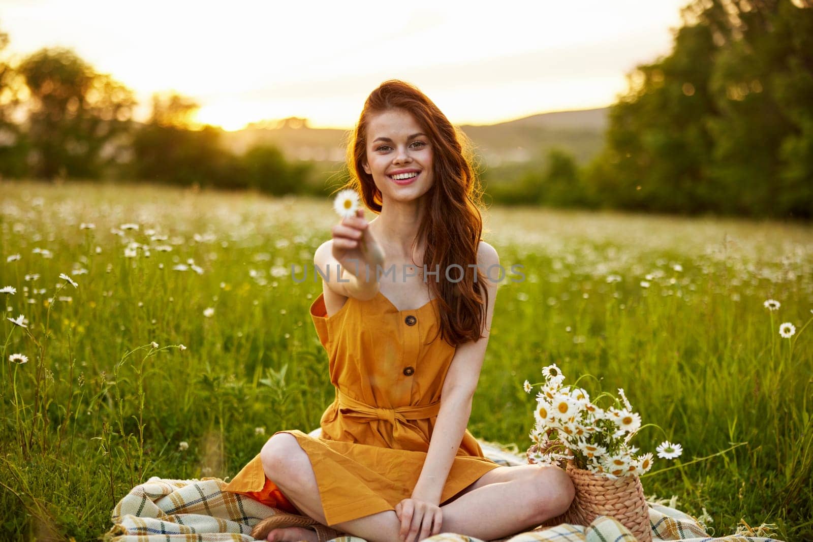 happy redhead woman sitting in a chamomile field on a plaid in a lotus position and holding a flower in her hands smiling at the camera. High quality photo