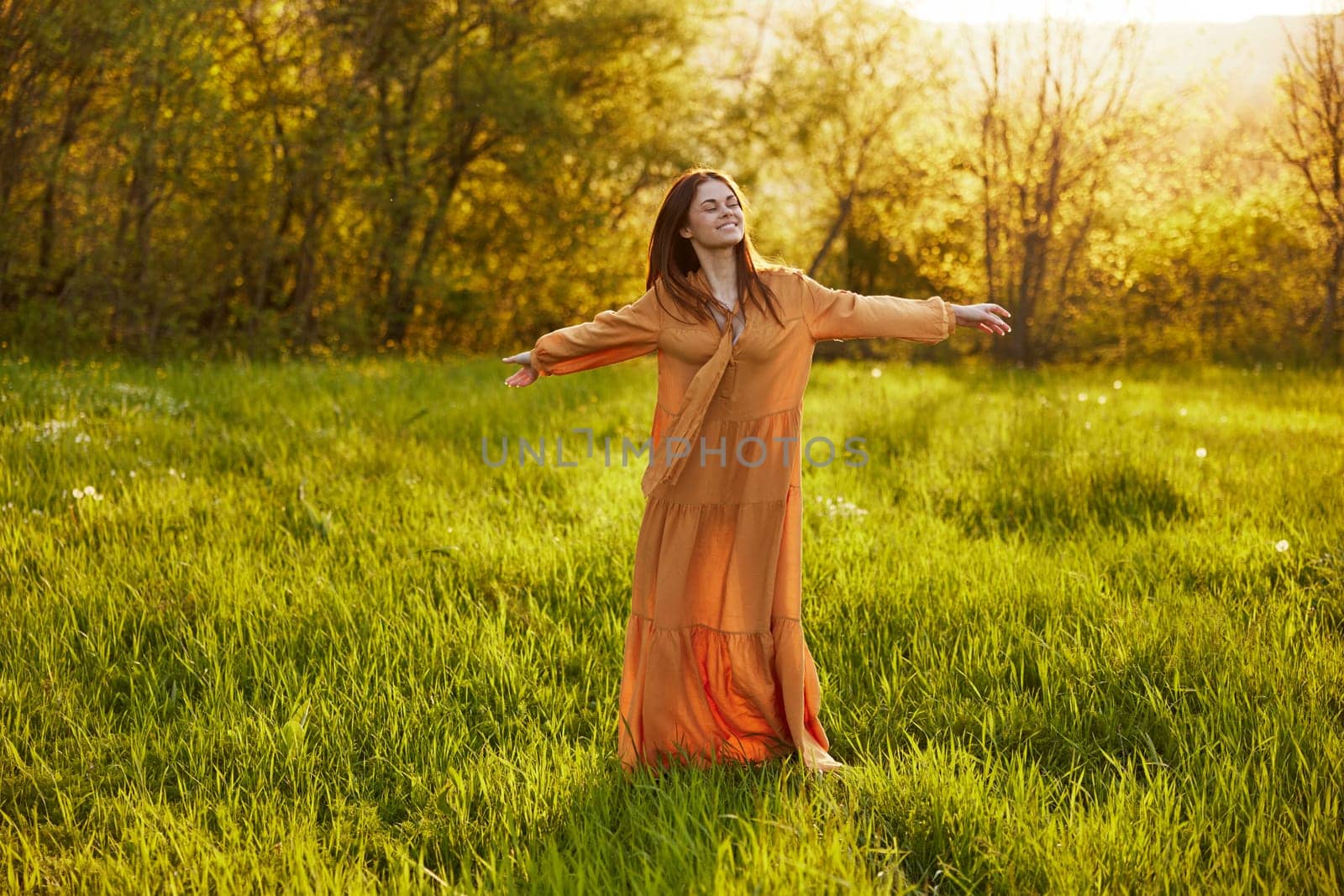 an attractive, slender, red-haired woman stands in a wide, green field during sunset in a long orange dress enjoying unity with nature and relaxation, raising her arms to the sides while facing the camera. High quality photo