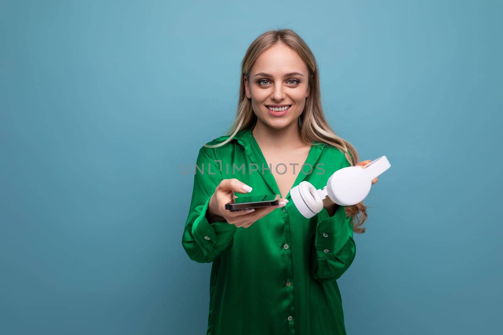 cute stylish attractive blonde girl with headphones and smartphone stretches out her hands on a blue background by TRMK