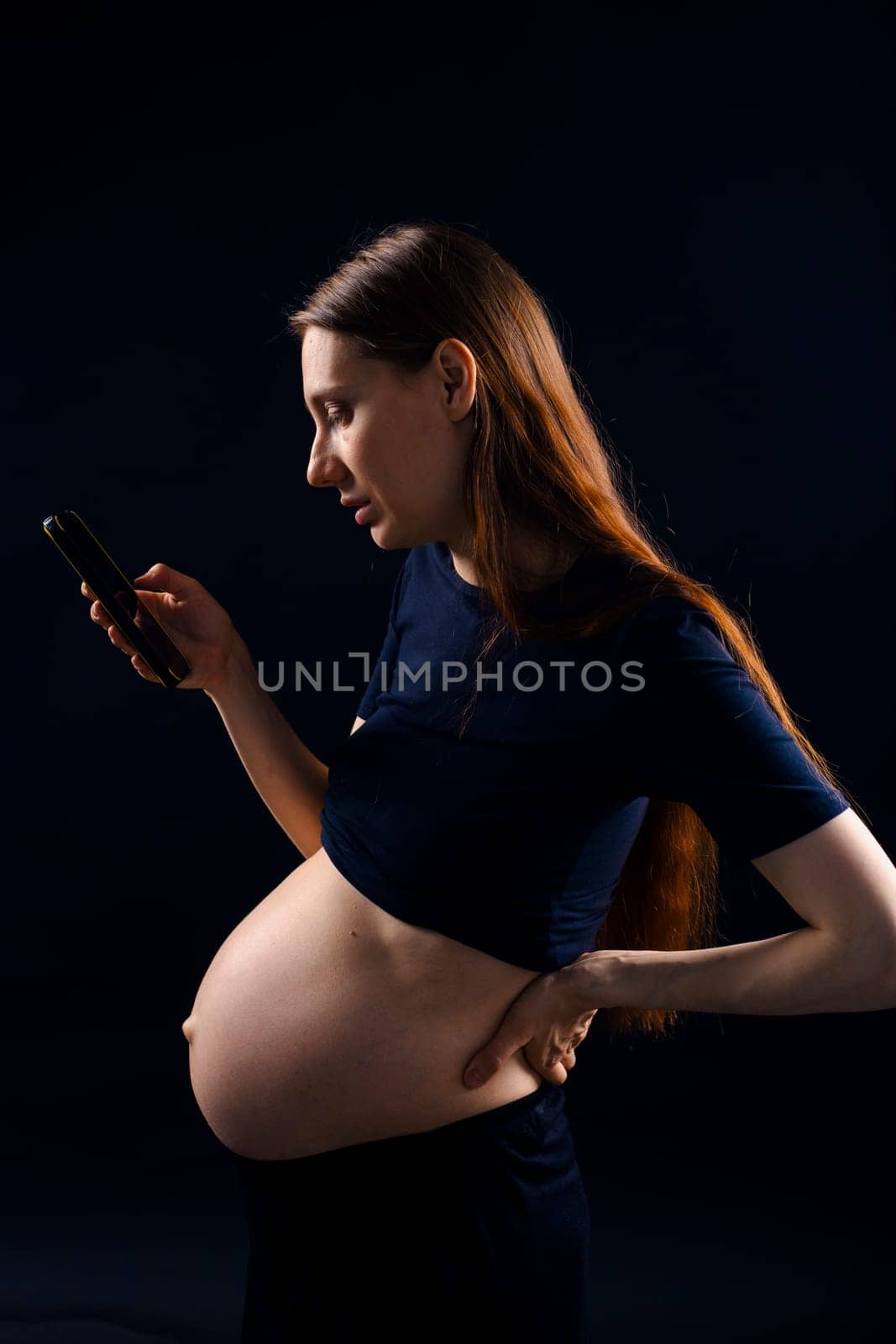 Beautiful pregnant woman with a smartphone over black background. Artistic low key light in studio. High quality photo