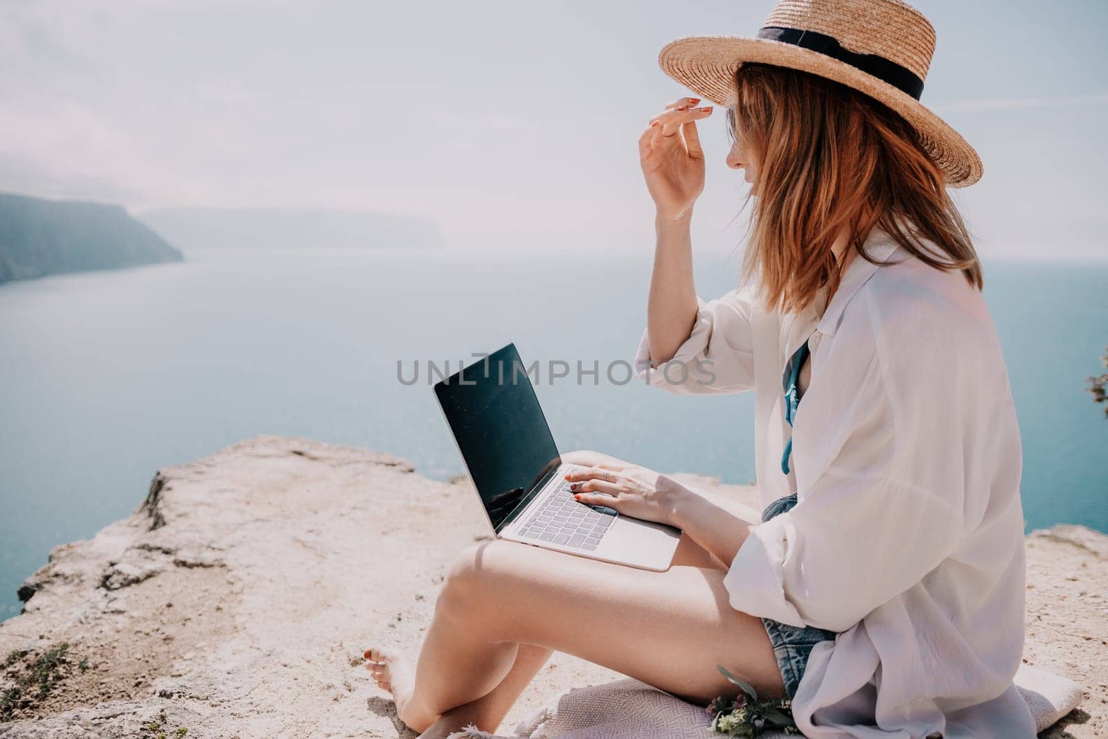 Successful business woman in yellow hat working on laptop by the sea. Pretty lady typing on computer at summer day outdoors. Freelance, travel and holidays concept.