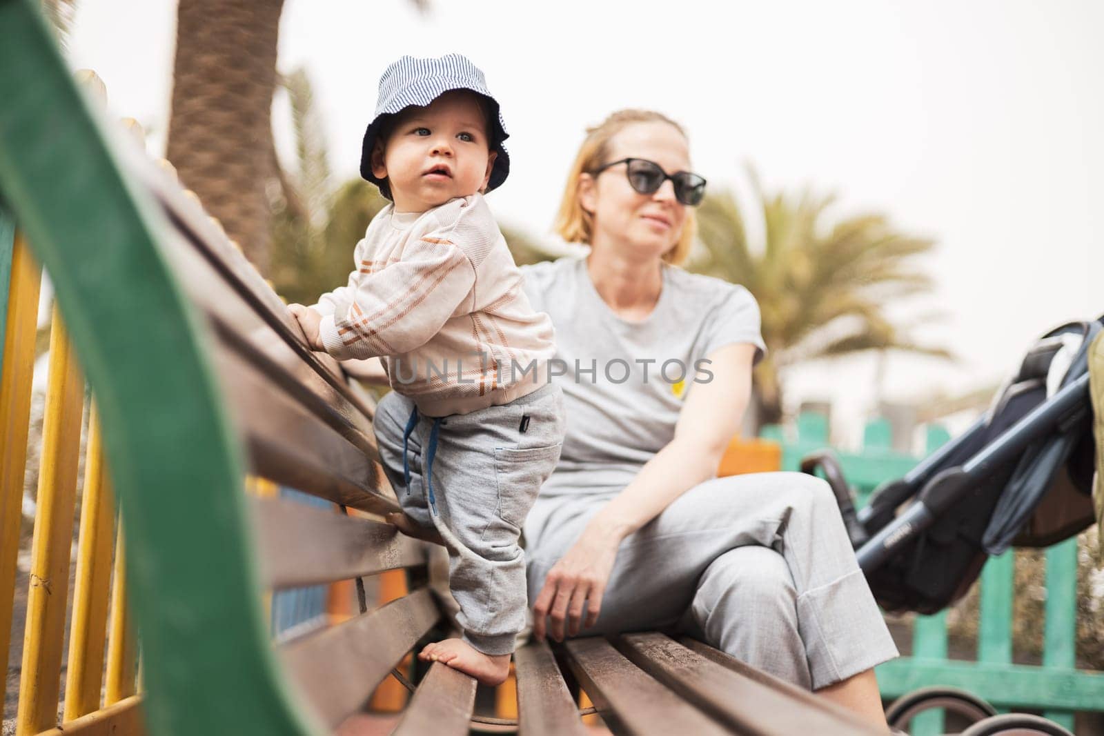 Young mother with her cute infant baby boy child on bench on urban children's playground on warm summer day. by kasto