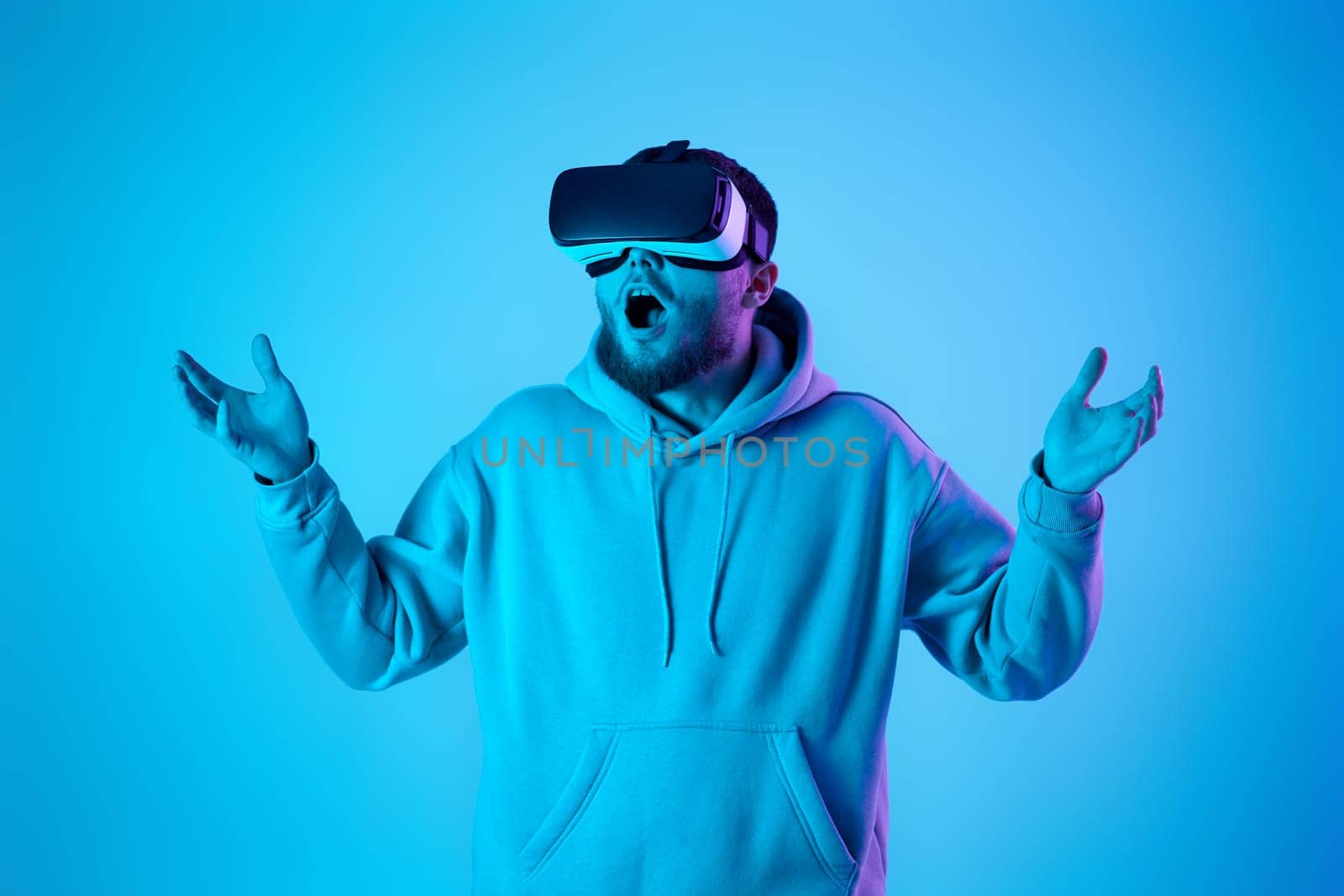 man in sweatshirt using virtual reality glasses and playing video games on blue background. Neon lighting