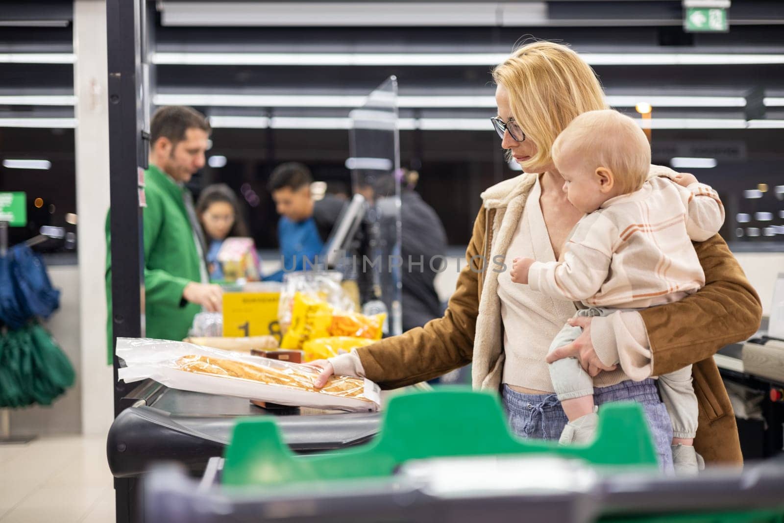Mother shopping with her infant baby boy, holding the child while stacking products at the cash register in supermarket grocery store. by kasto