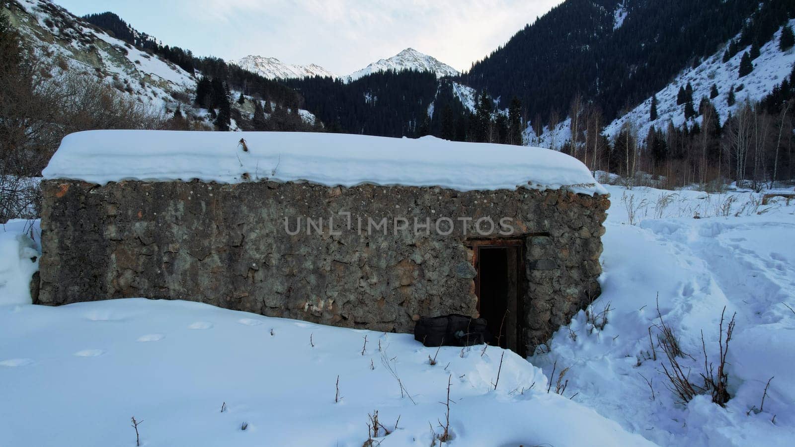 An old stone prison or hut in the mountains. Snowy mountains, forest and clouds against a blue sky. Tall dry bushes peek out from under the snow. Birch and spruce trees are visible in the distance.