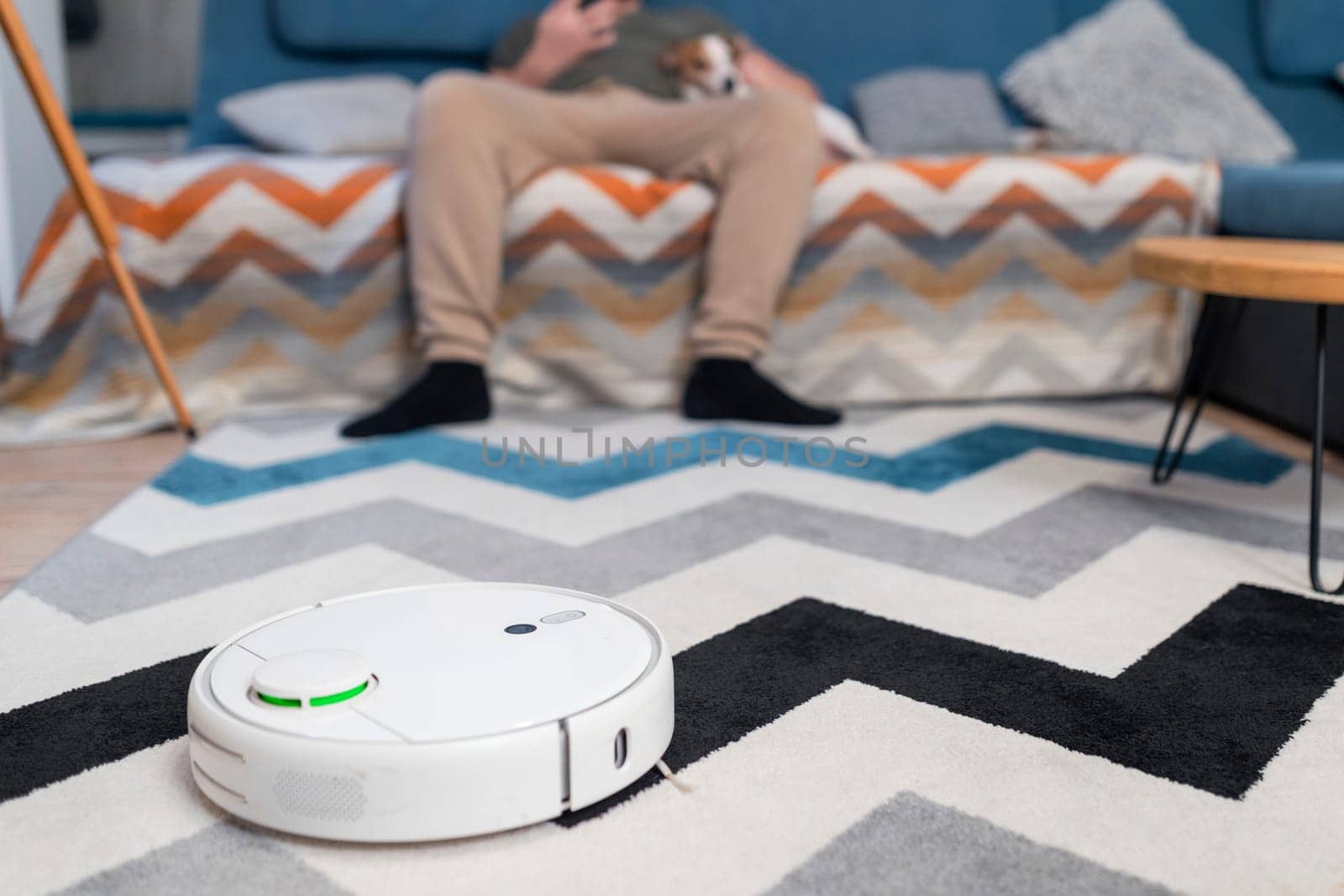 Man couple sitting on sofa with dogs cleaning floor using vacuum robot at home.