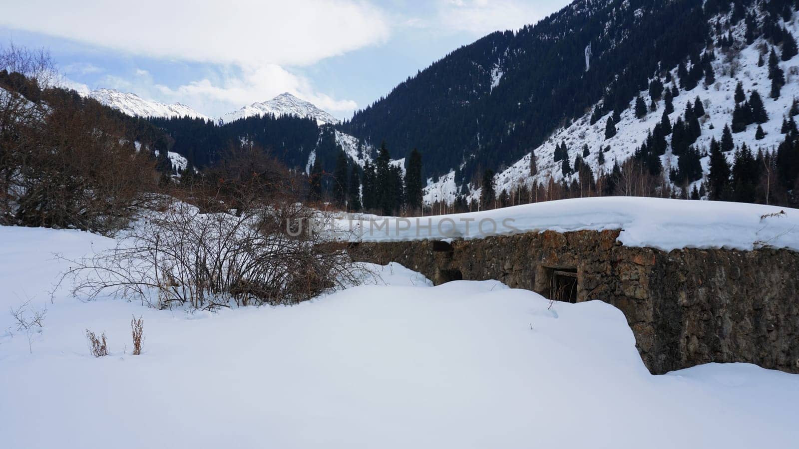 An old stone prison or hut in the mountains. Snowy mountains, forest and clouds against a blue sky. Tall dry bushes peek out from under the snow. Birch and spruce trees are visible in the distance.