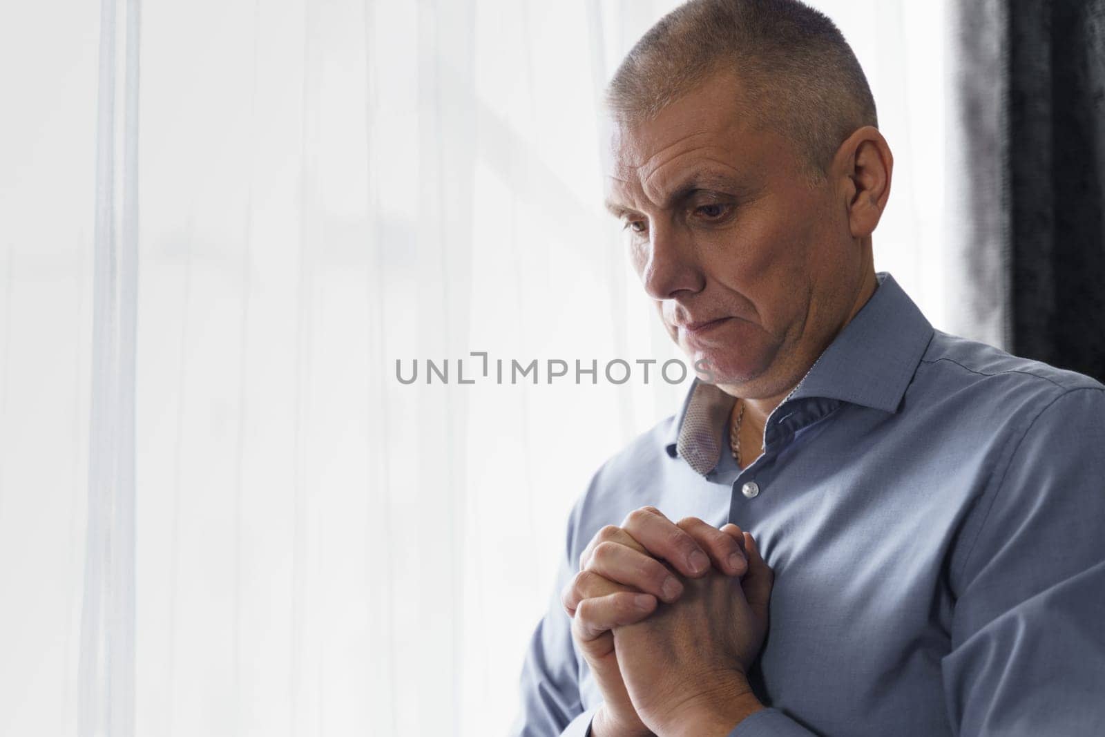 Portrait of a man who stands at the window clenched his hands in front of him, concentrated, prays or thinks. Side view.