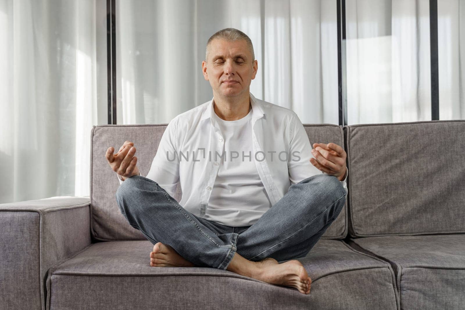 Portrait of a man who sits on a sofa in a room in a lotus position, meditates with his eyes closed. The man is doing yoga.