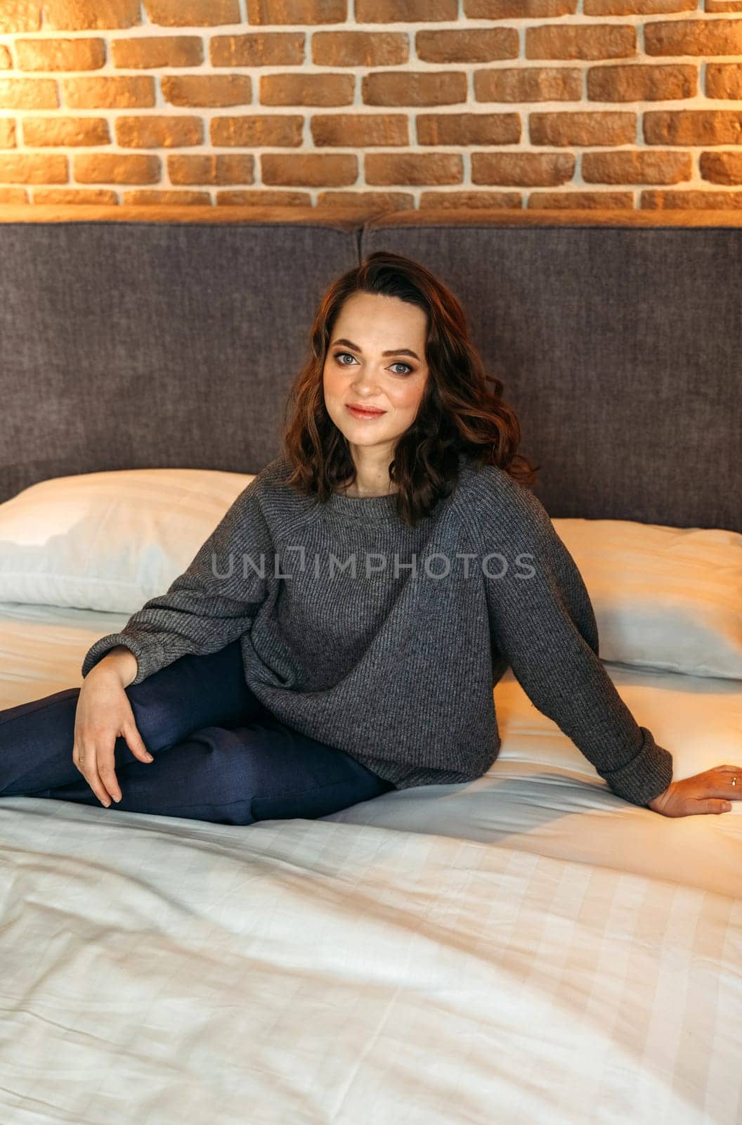 An attractive woman is sitting on a bed in a bedroom wearing blue trousers and a gray jumper. Smiling and looking at the camera. Vertical frame.