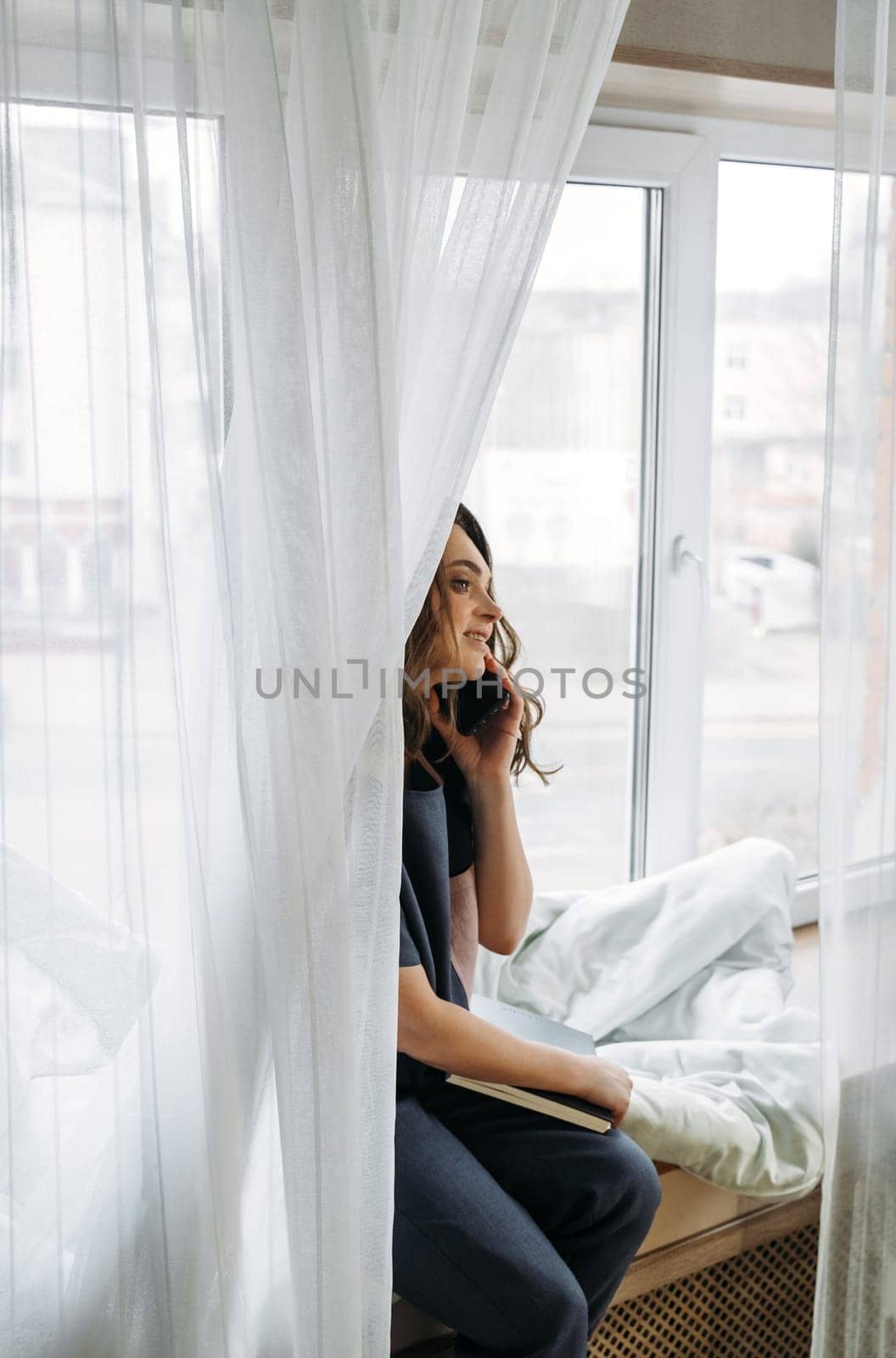 A young woman sitting on a windowsill talking to clients, holding a notepad in her hands. Vertical frame.